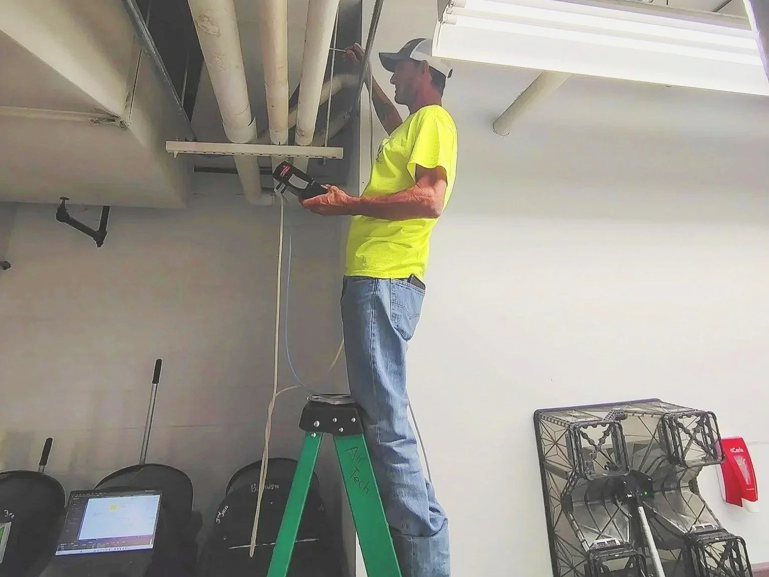 A man standing on a green ladder working on ceiling pipes with a black device in hand, in an indoor setting.