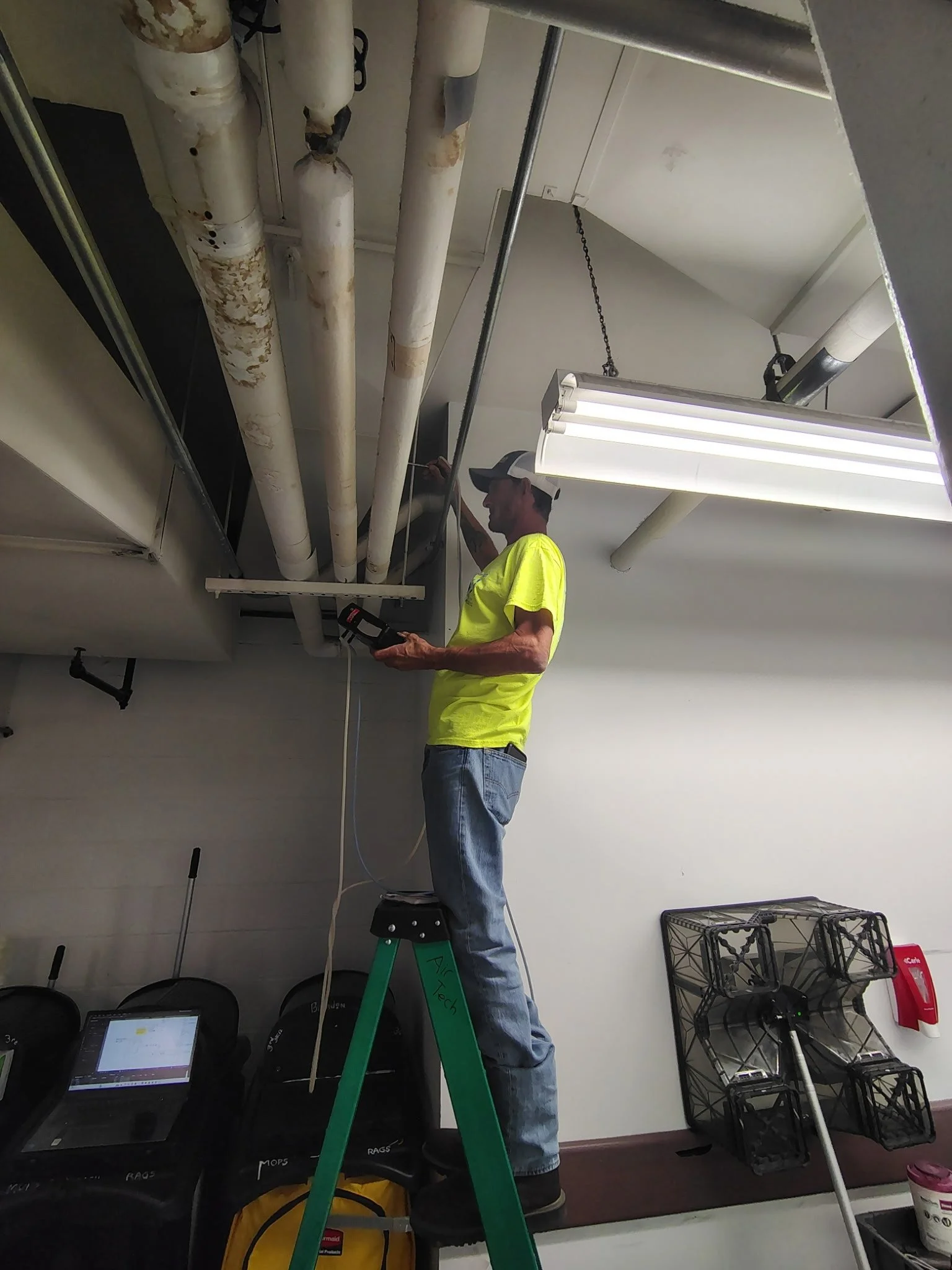 A technician standing on a step stool repairing or inspecting pipes on a ceiling using a tool. The room has industrial pipes, a fluorescent light fixture, and some equipment or storage on the floor.