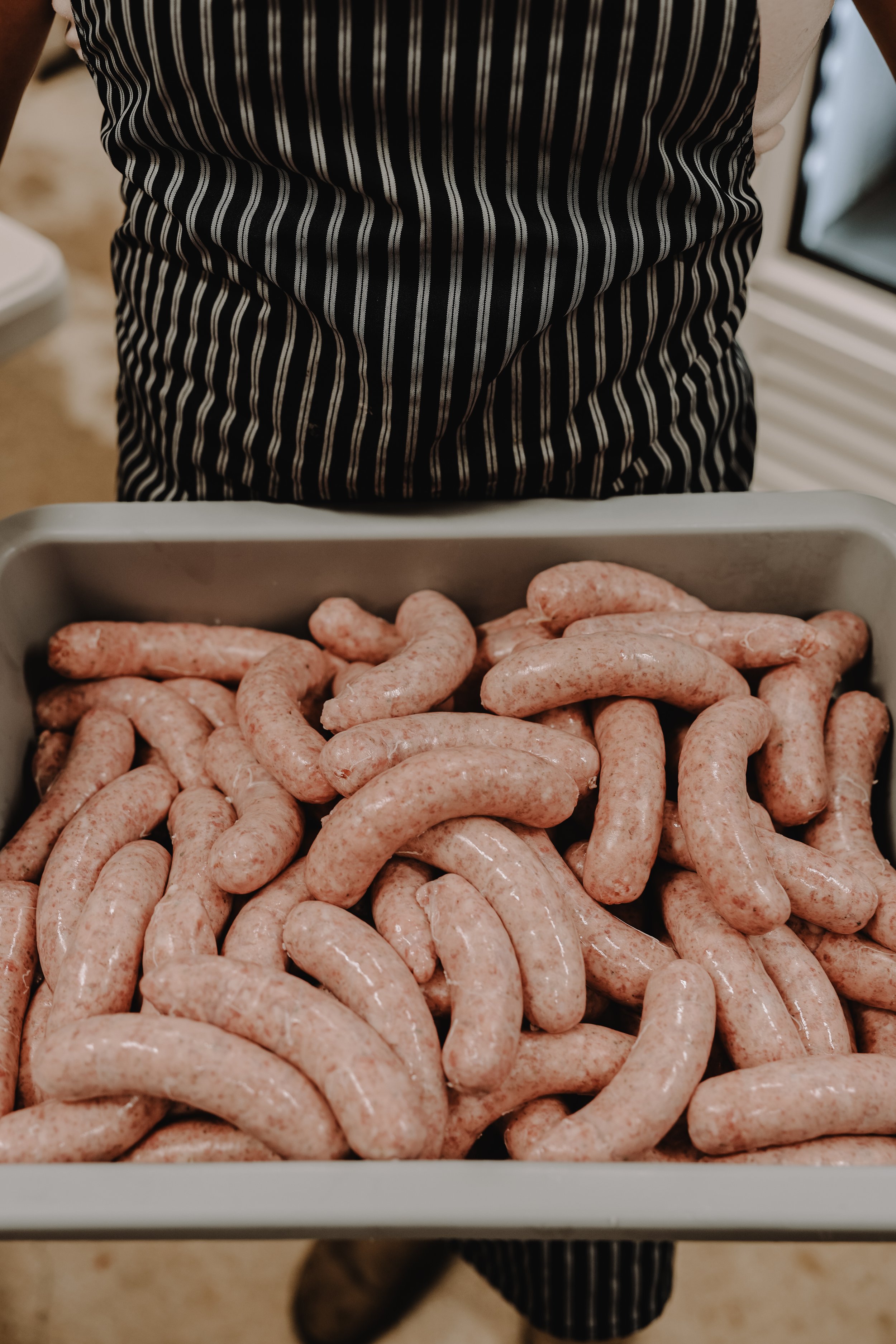 A person wearing a black and white striped apron holding a tray full of raw sausages.