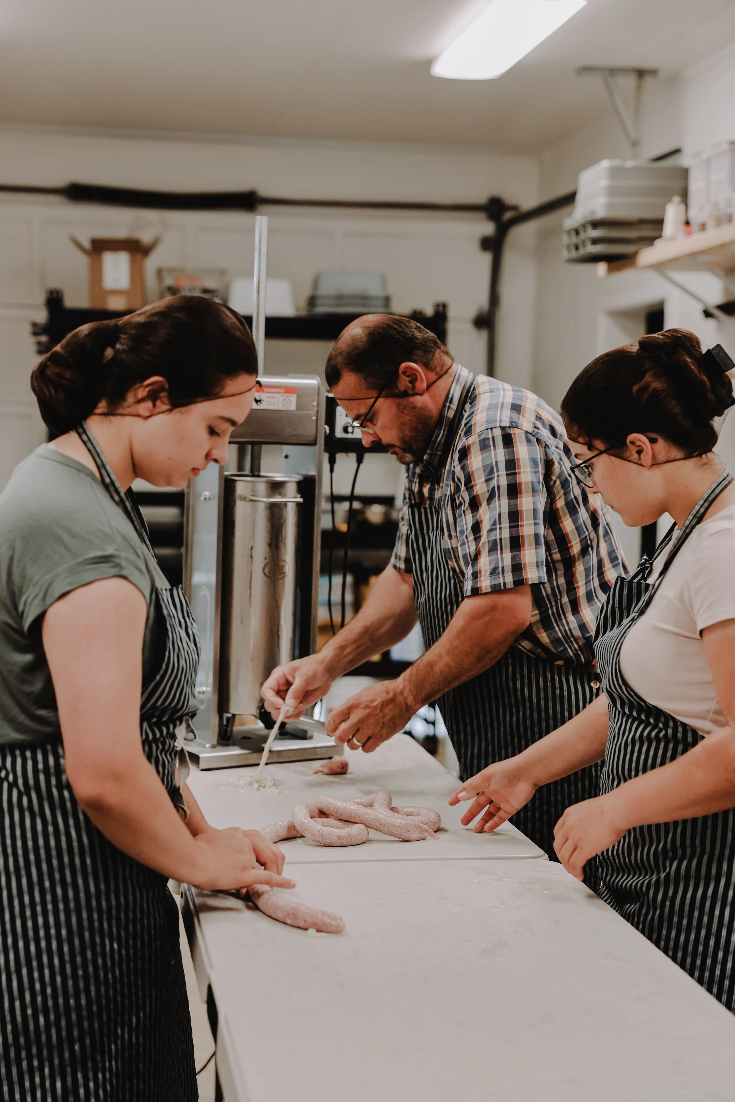 Three people working together in a kitchen, preparing sausages on a counter.
