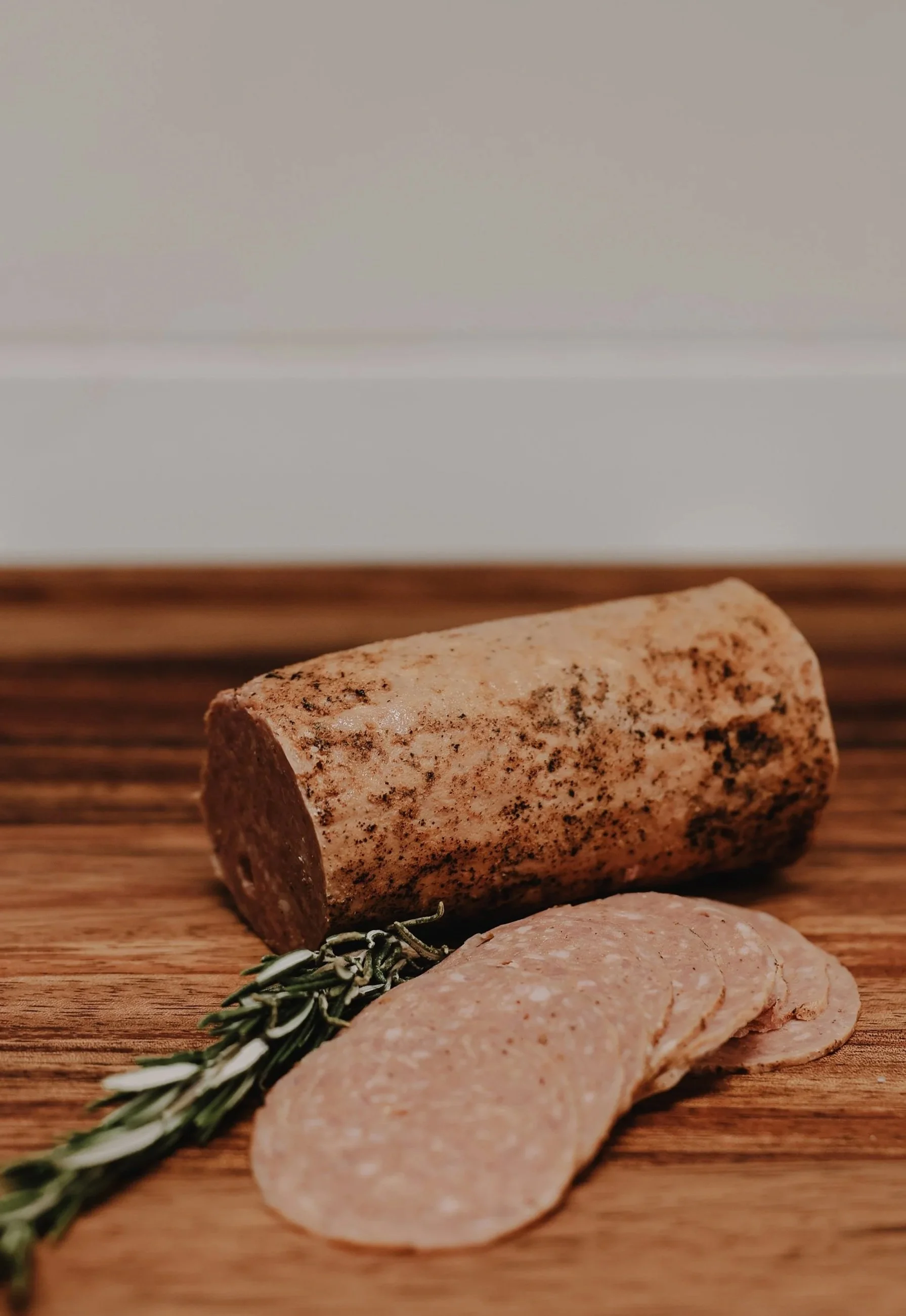 Sliced cured meat, a piece of salami, garnished with a sprig of rosemary, on a wooden cutting board.