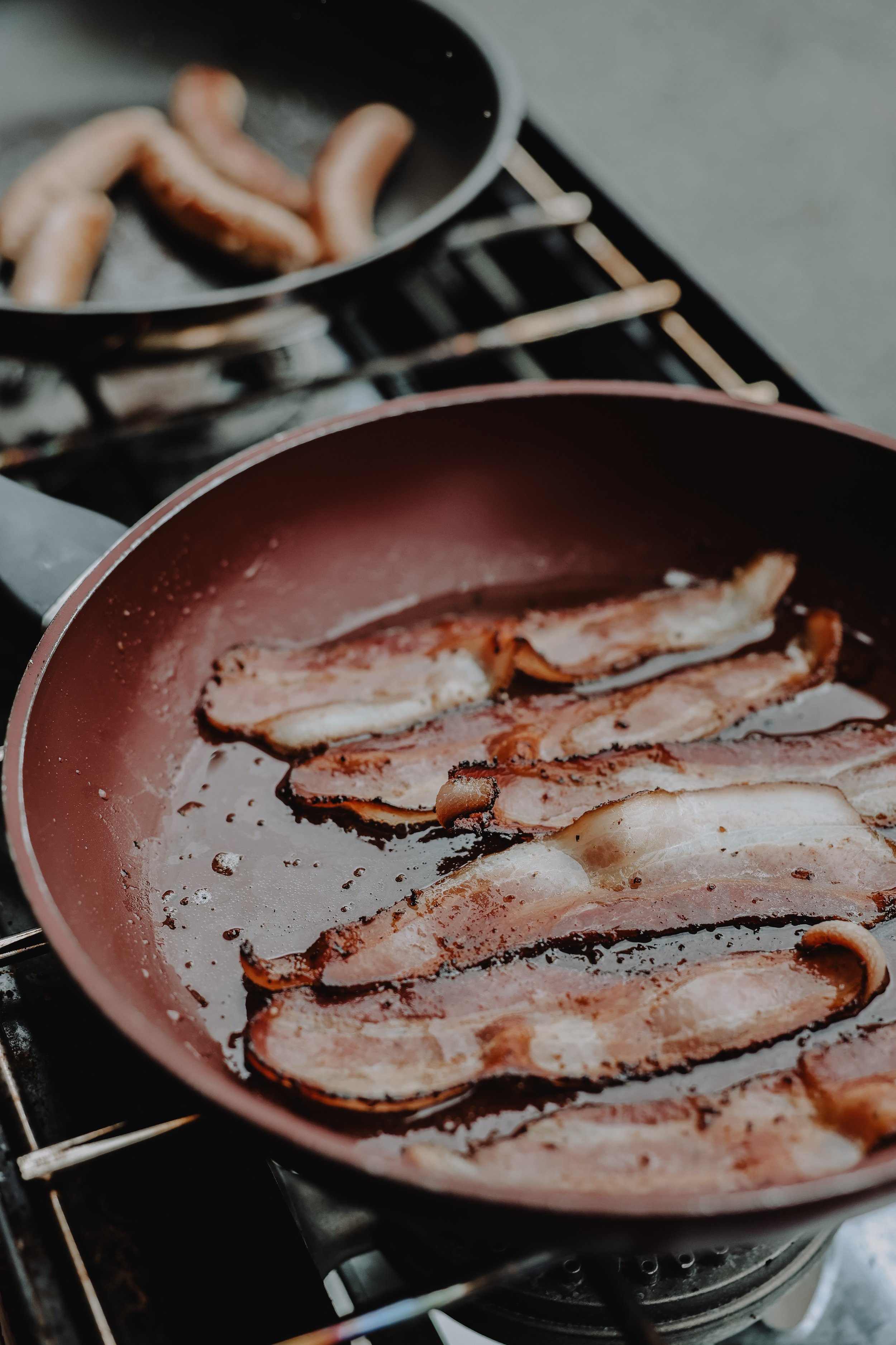 Bacon frying in a pink skillet on a stove, with sausage links cooking in a black pan in the background.