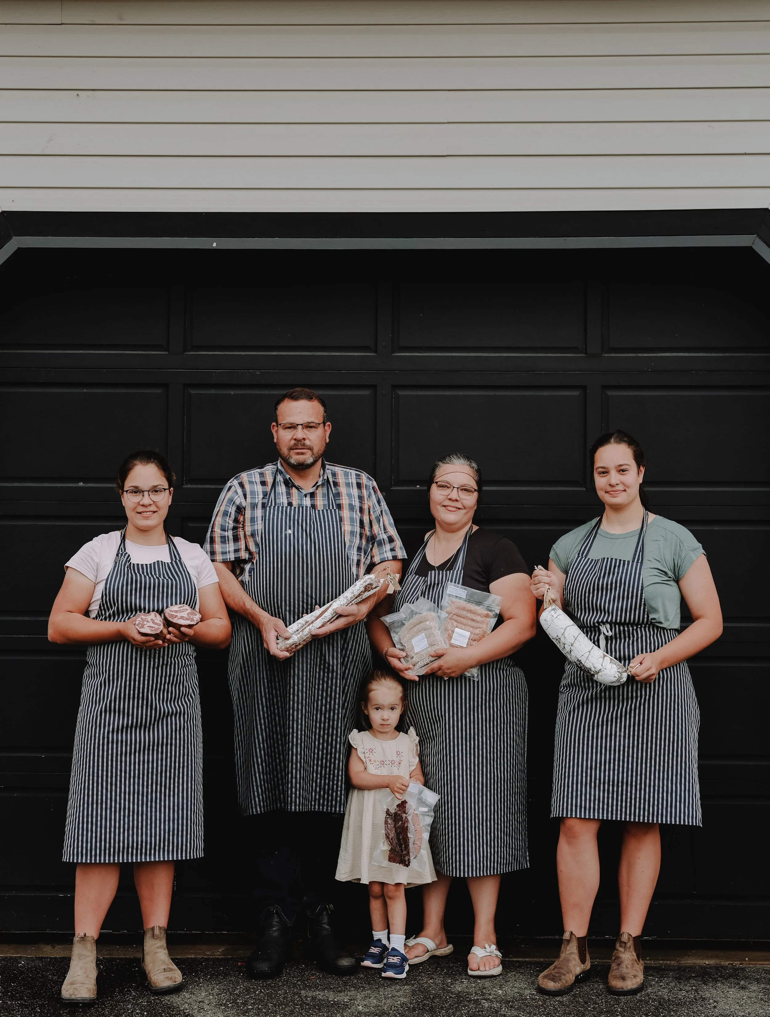 Group of five people, including two children, standing in front of a black garage door, wearing striped aprons and holding various types of meat products.