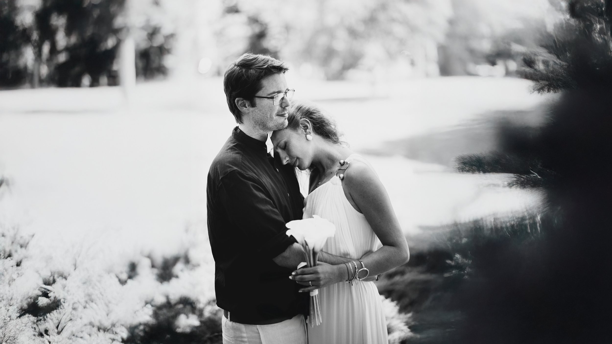 A black and white photograph of a couple embracing outdoors, with the woman resting her head on the man's shoulder while holding a bouquet of calla lilies, against a blurred natural background.