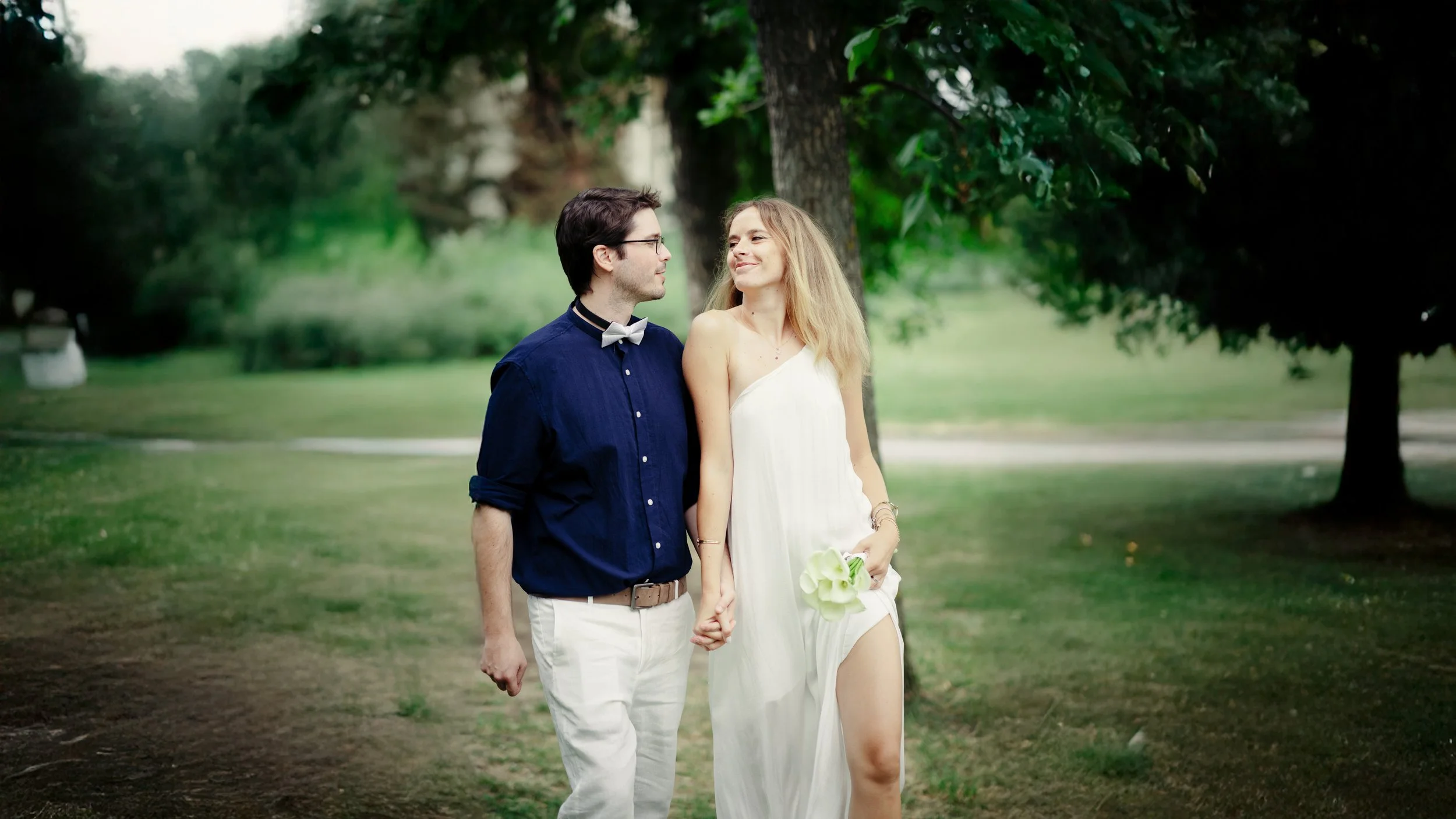 A young couple is holding hands and walking through a park with green grass and trees; the woman is wearing a white dress and holding a bouquet, and the man is dressed in a navy shirt with a bowtie.