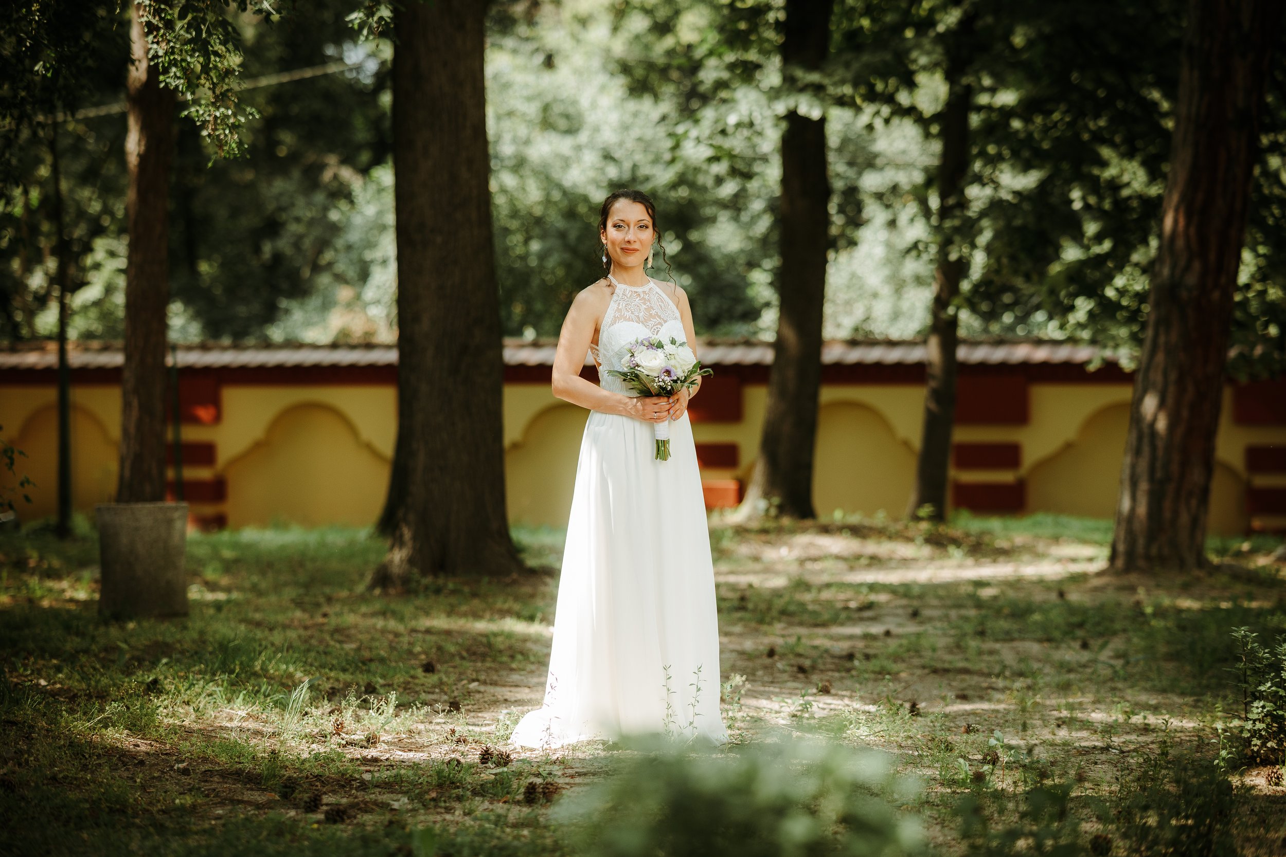 A bride standing outdoors in a wooded area, wearing a white wedding dress with lace details, holding a bouquet of flowers, with trees and a decorative wall in the background.