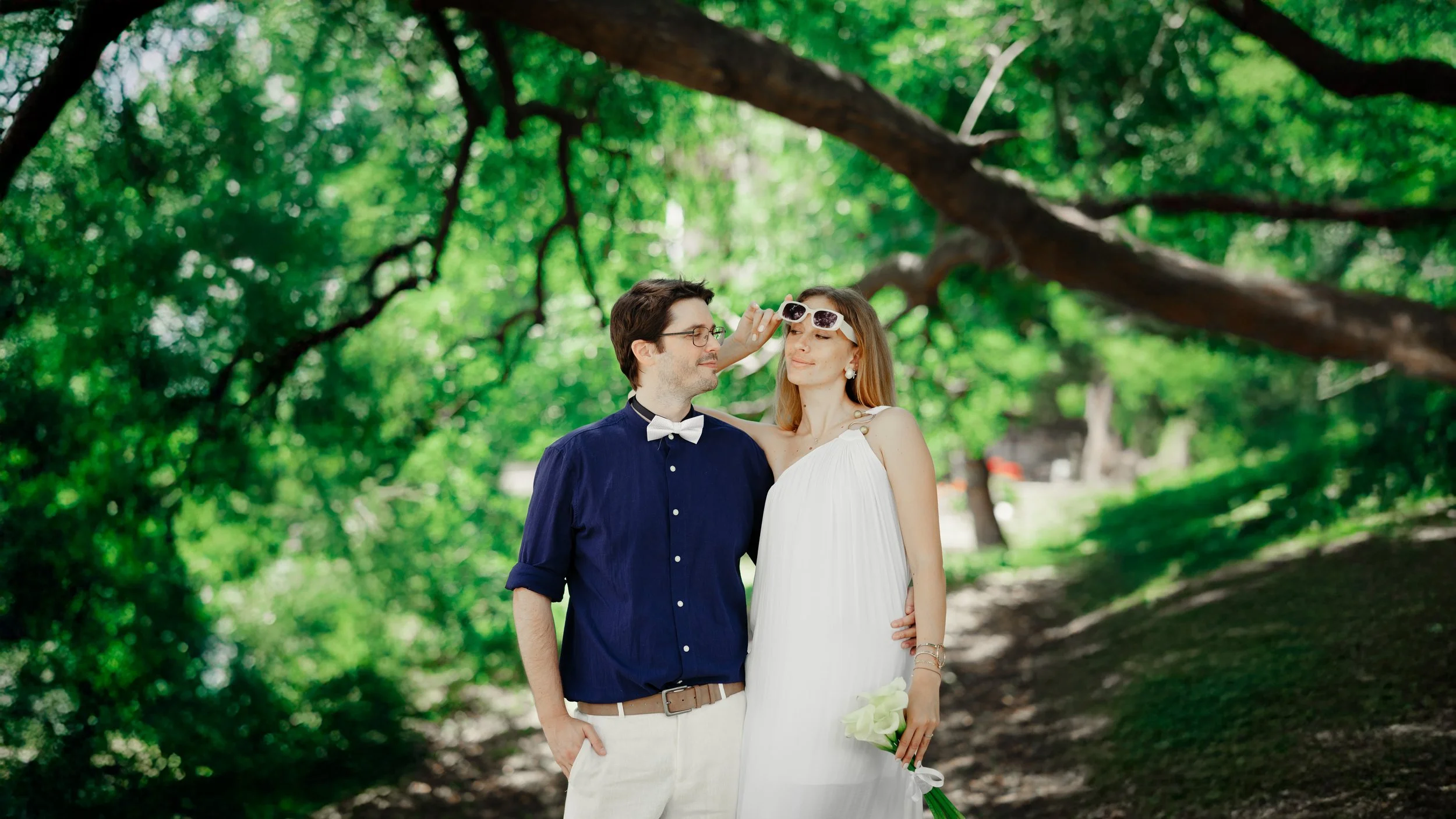 A couple in wedding attire standing outdoors under a large tree with green leaves. The woman is holding a bouquet of white flowers and wearing sunglasses, which the man is adjusting. The man is dressed in a navy shirt, white pants, and a bow tie.