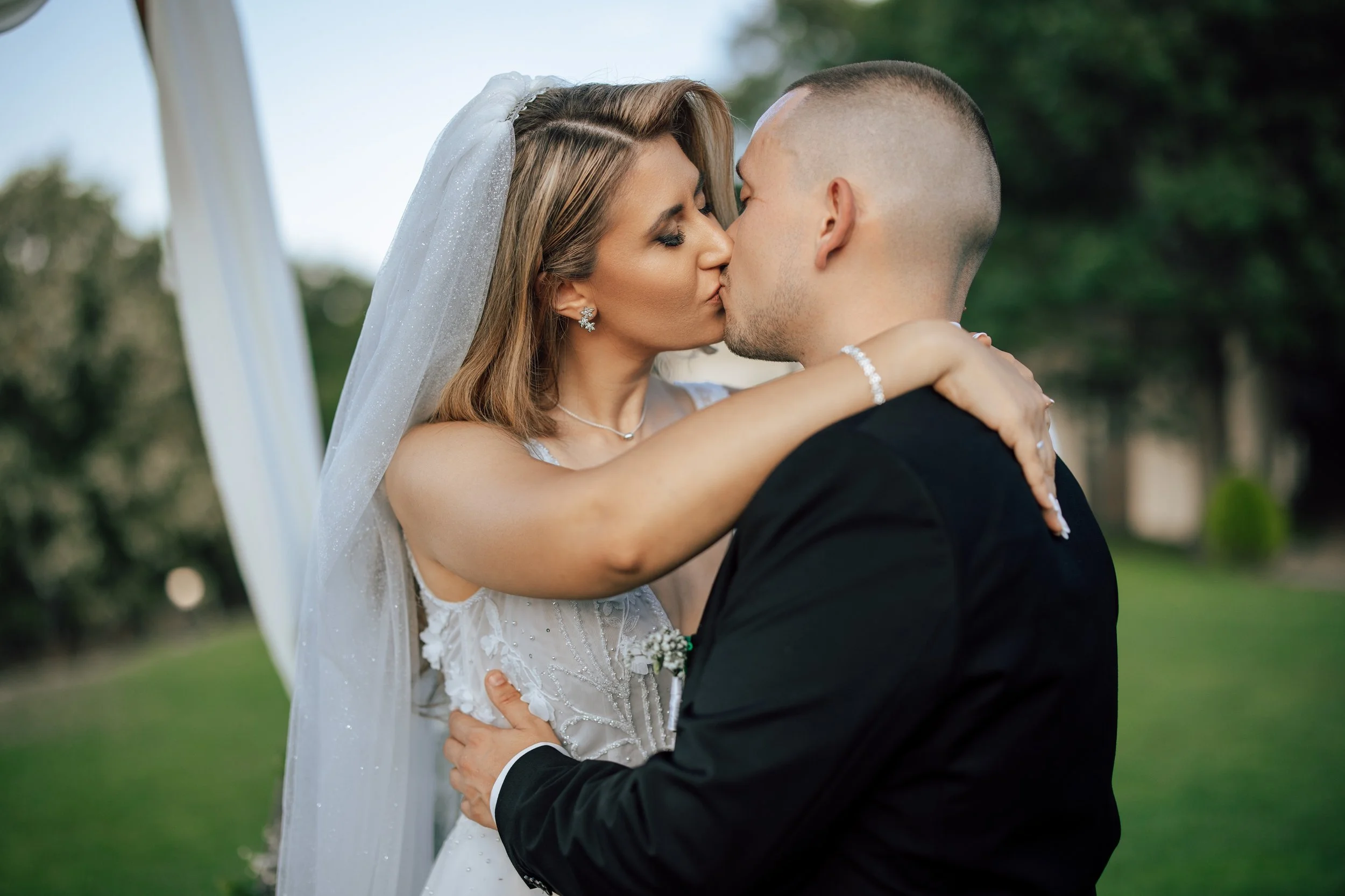A bride and groom kissing outdoors with greenery in the background.