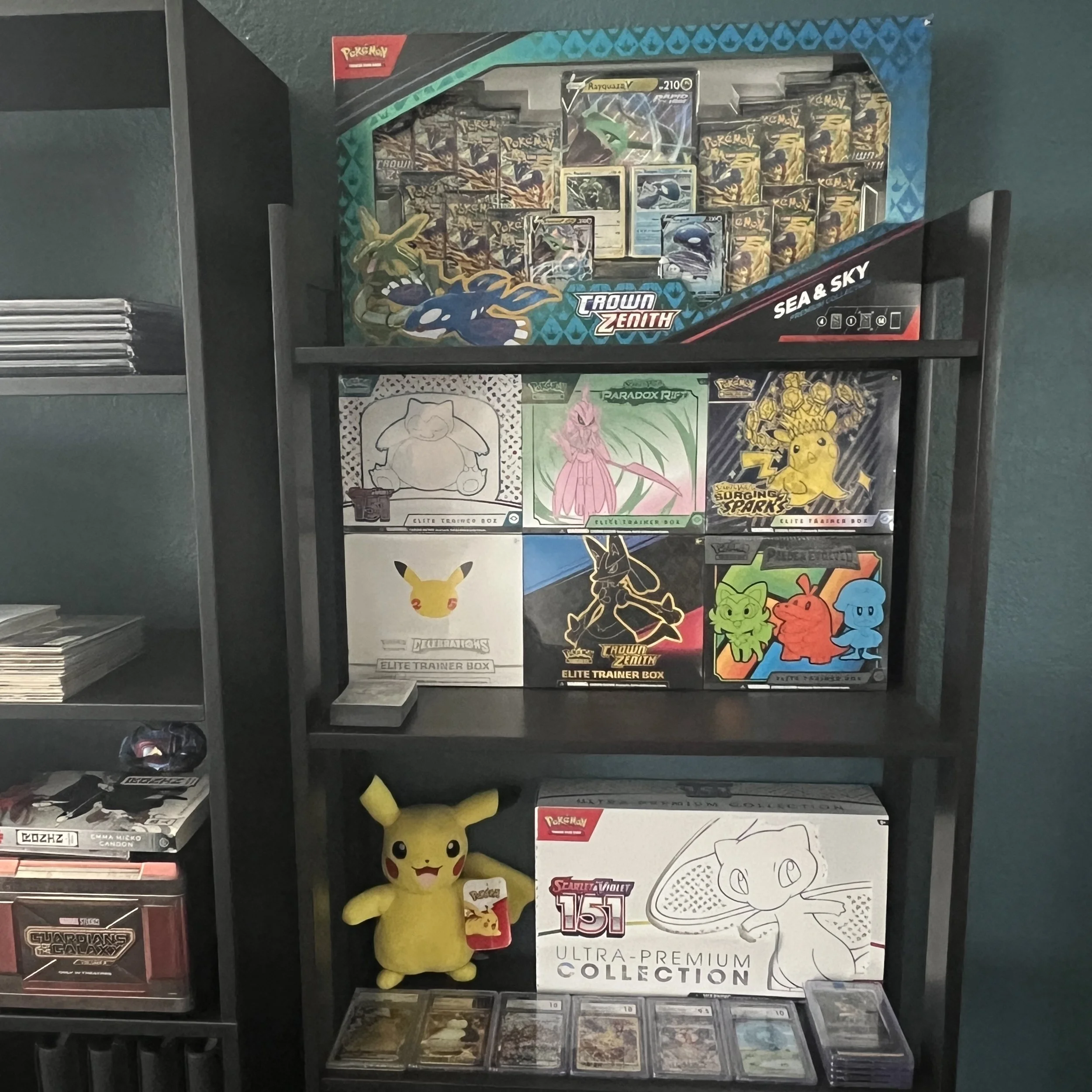 A black shelving unit displaying Pokémon trading cards, plush Pikachu, and Pokémon merchandise, including a large Pokémon card collection box on the lower shelf.