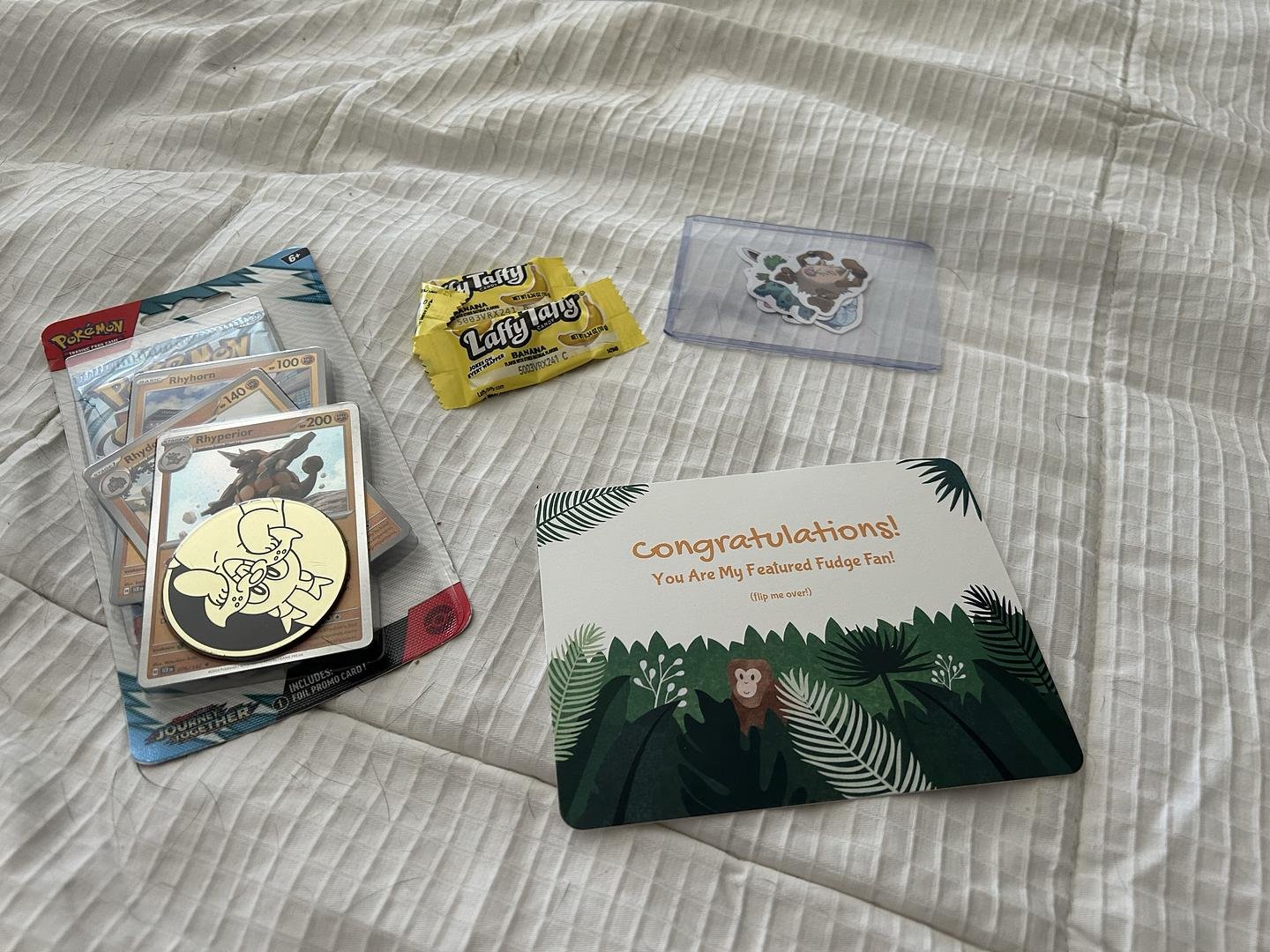 A collection of Pokémon trading cards, a yellow Laffy Taffy banana-flavored candy, a clear plastic slot with Pokémon stickers, and a congratulatory card on a white textured bedspread.