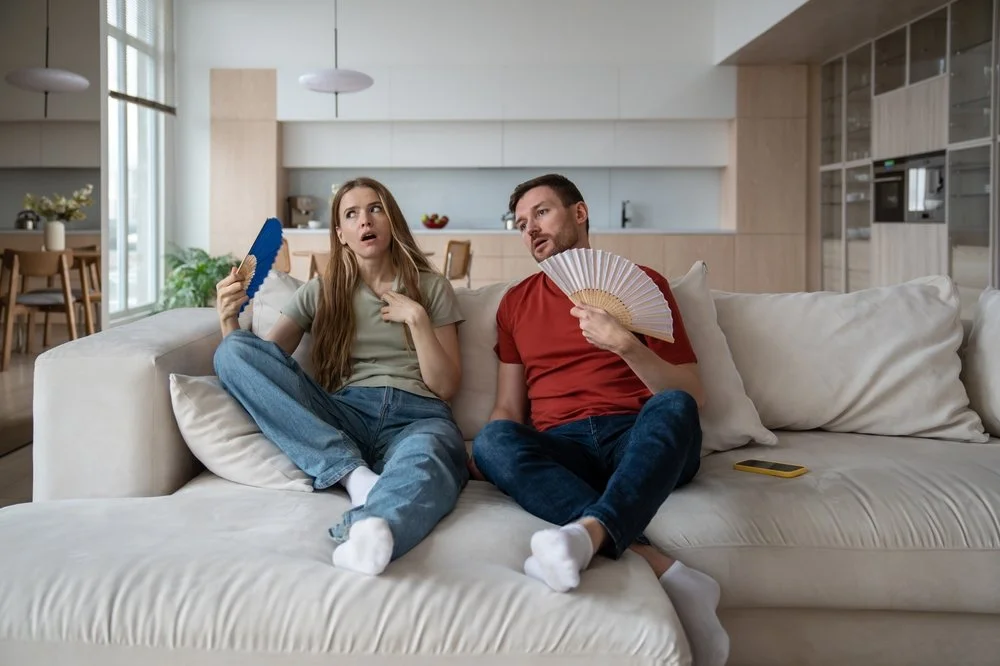 A young woman and a man sitting on a white sofa in a modern, well-lit kitchen and living space. The woman appears surprised or confused, holding a blue fan, while the man is holding a white fan with a wooden handle. There is a yellow smartphone on the sofa beside them.