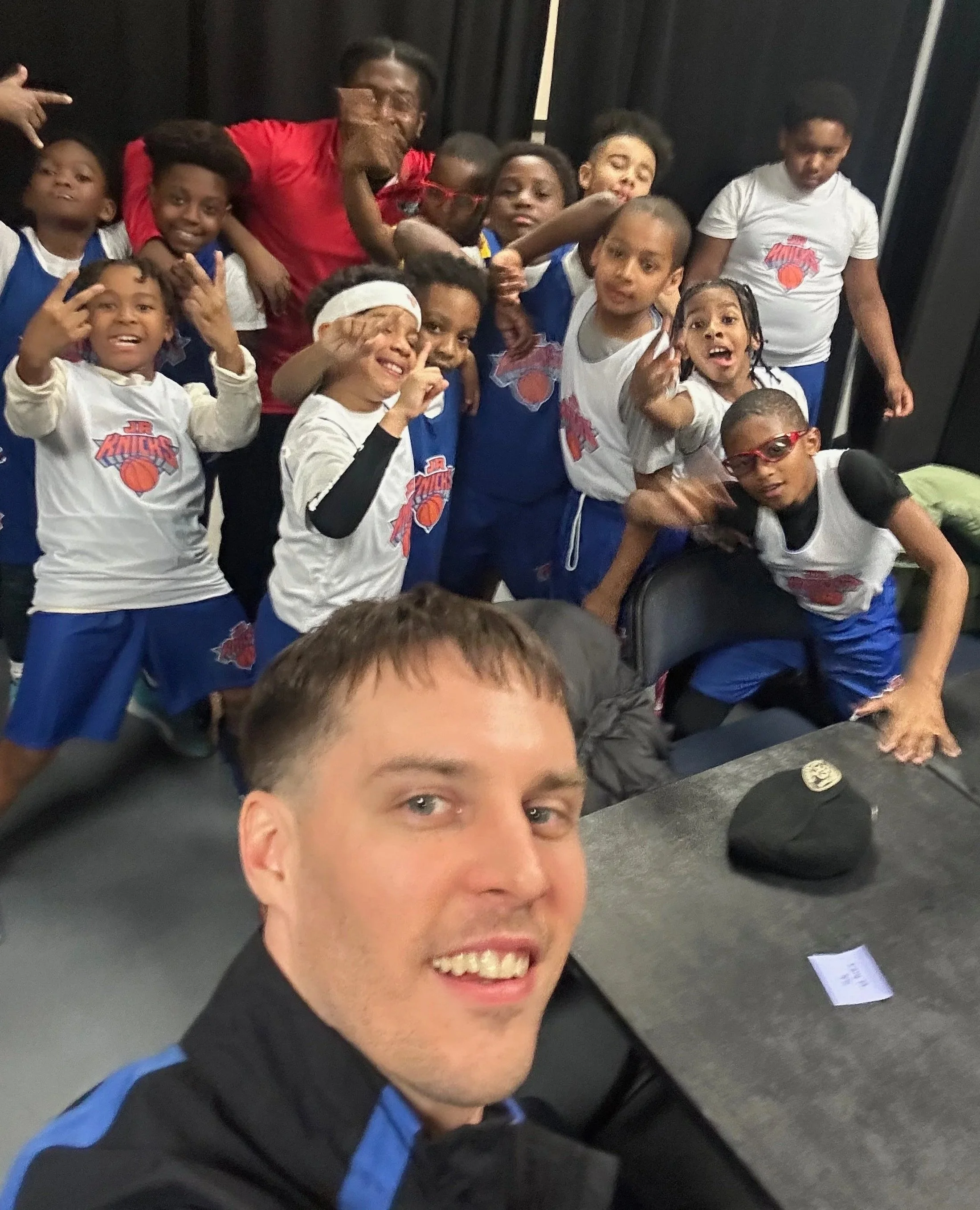 A group of young basketball players and two adults posing for a selfie in a gym. The kids are wearing basketball jerseys and making various gestures, smiling and excited, with black curtains in the background.