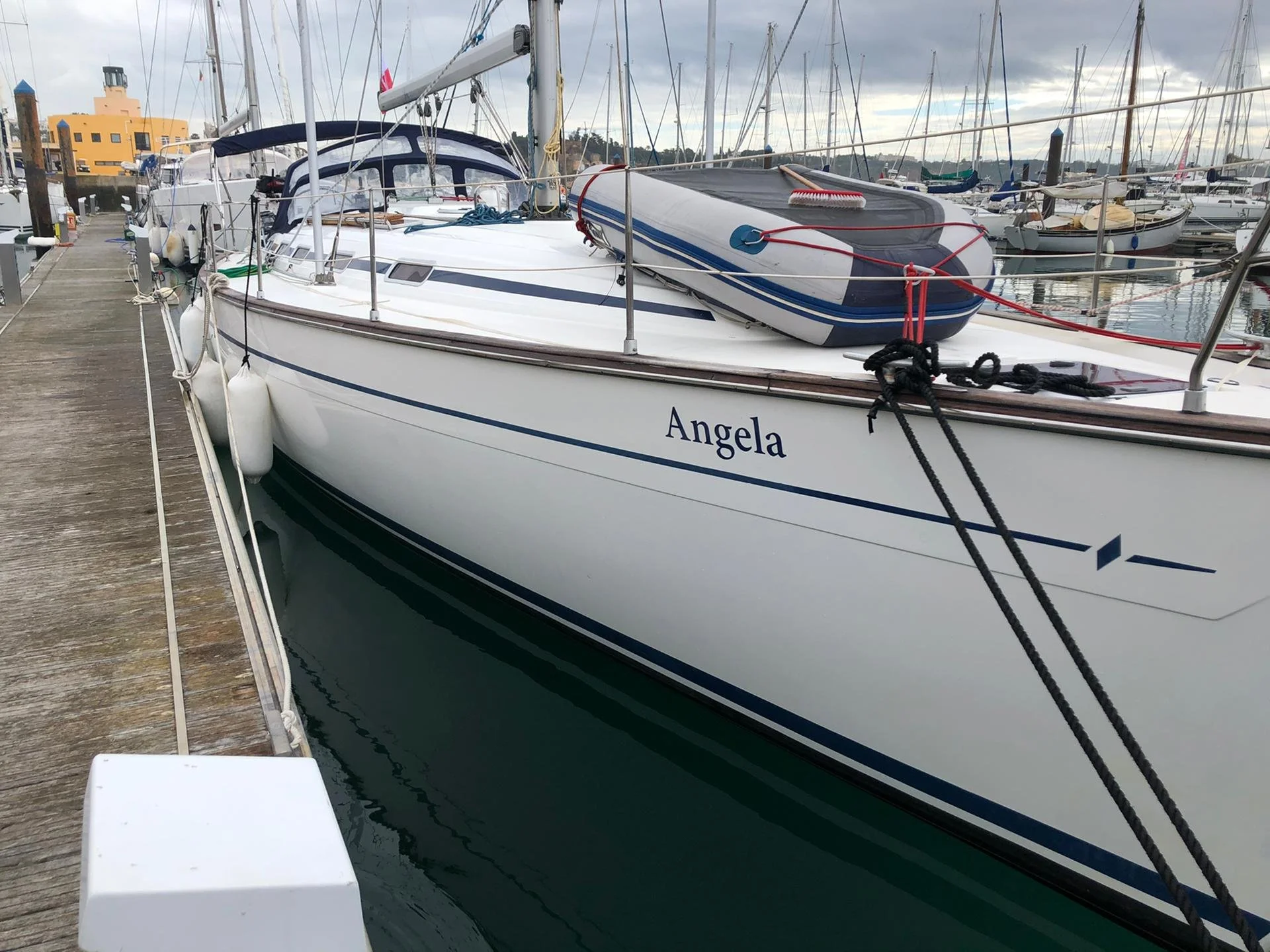 A white sailboat named Angela docked at a marina with other sailboats in the background and cloudy sky overhead.