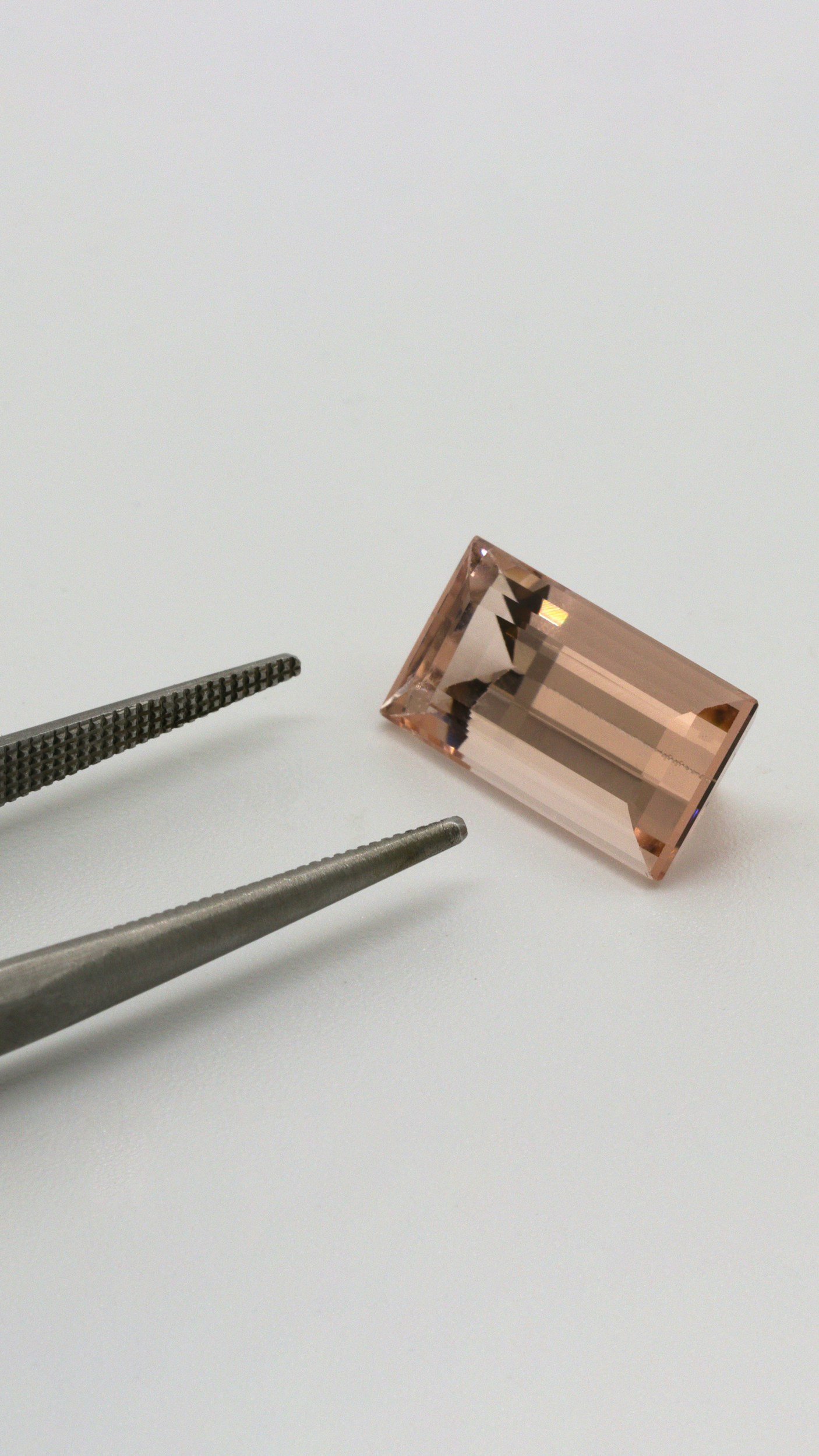 A large rectangular pink gemstone, likely a morganite, is placed on a white surface with a pair of metal tweezers nearby.