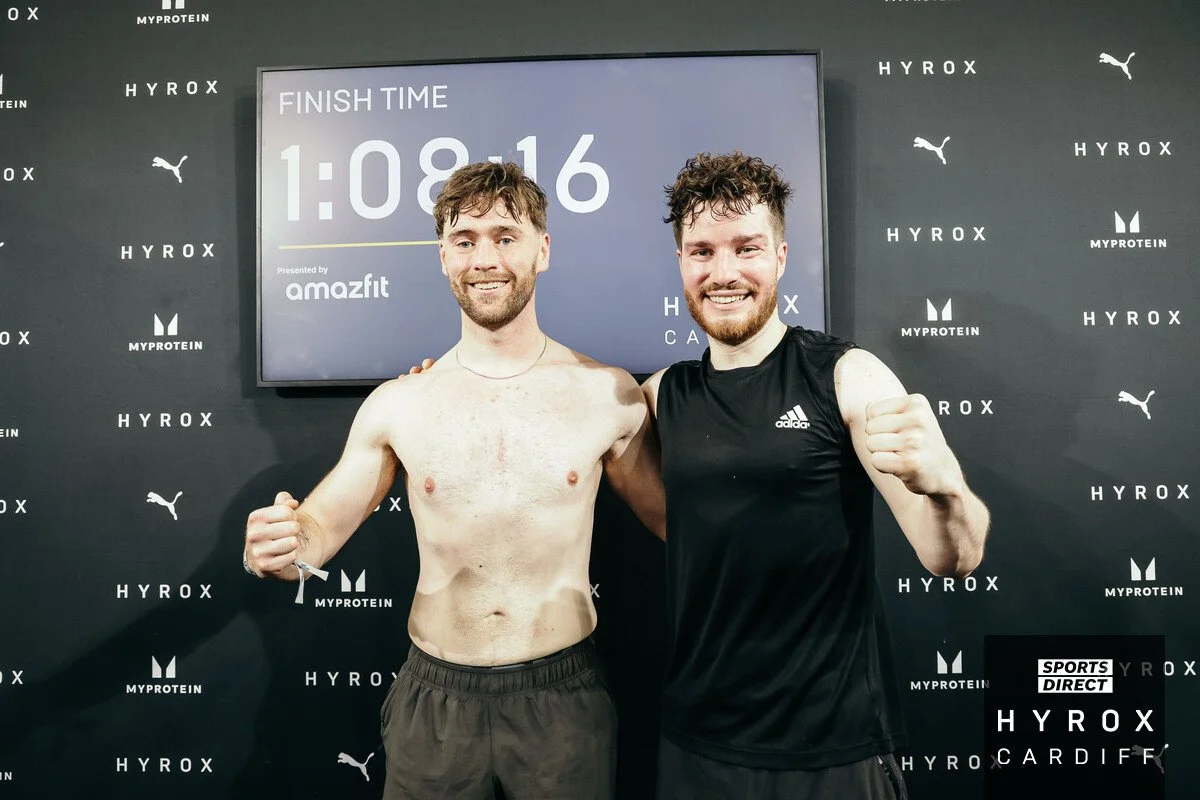 Two male athletes celebrating after a race, posing with fists raised, in front of a backdrop with logos including BT, MYPROTEIN, HYROX, Puma, and SPORTS DIRECT. One is shirtless, and the other is wearing a black sports shirt.