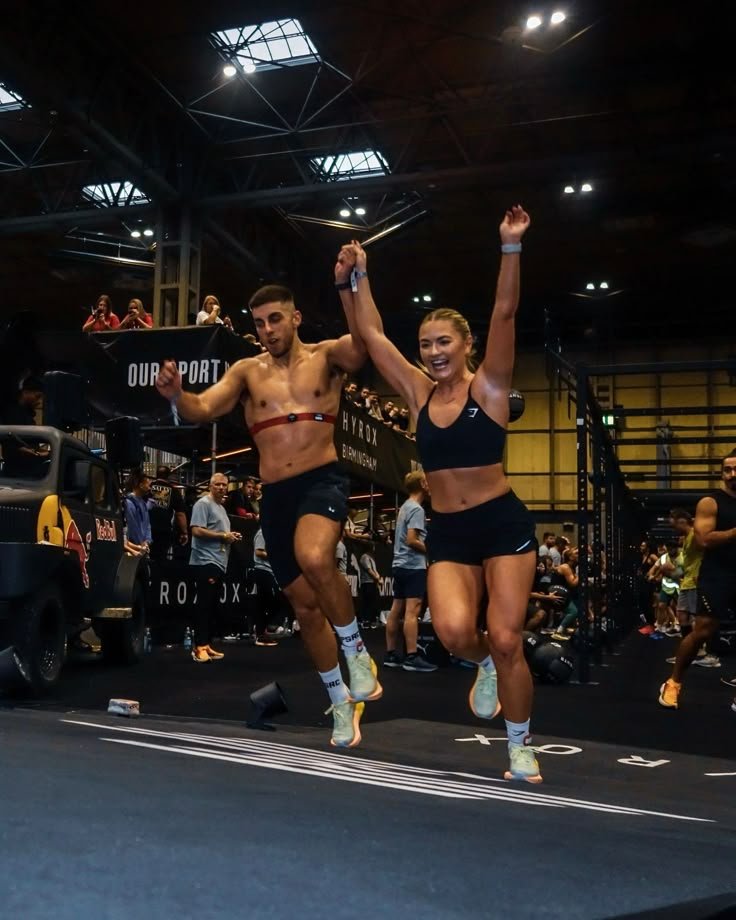 Two athletes celebrating after finishing a race, holding hands and jumping in an indoor sports arena with an audience in the background.