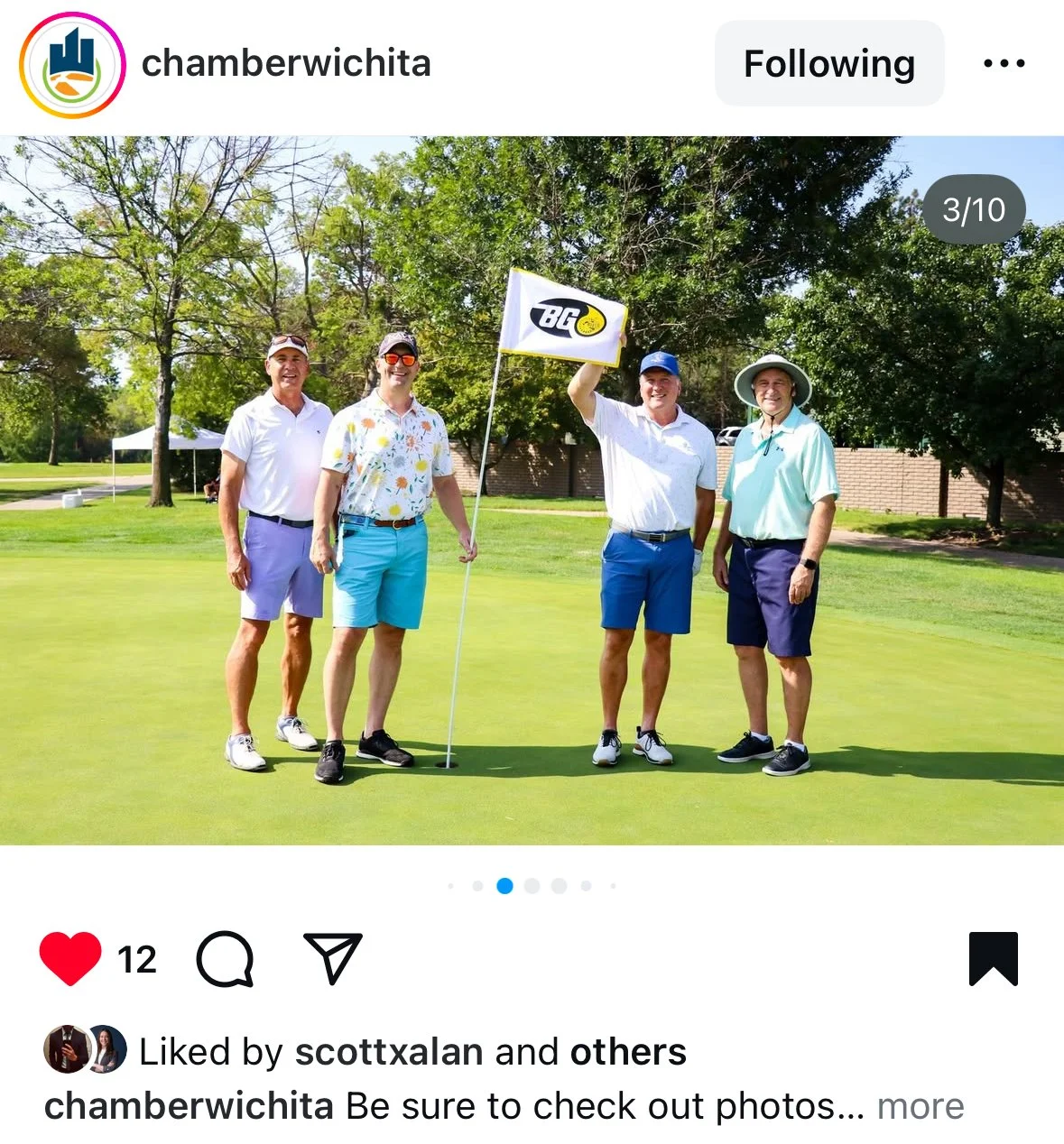 Four men standing on a golf course, one holding a flag with a logo, smiling at the camera on a sunny day with trees in the background.