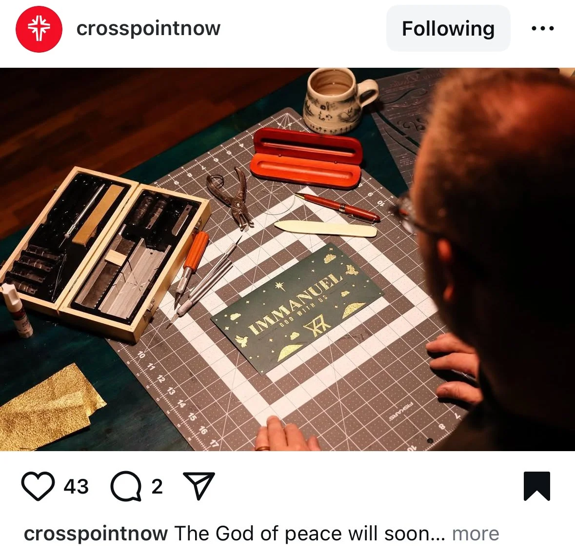 A person working on a holiday-themed greeting card that reads 'IMMANUEL GOD WITH US' surrounded by crafting tools, scissors, pens, a mug, and a crafting mat.