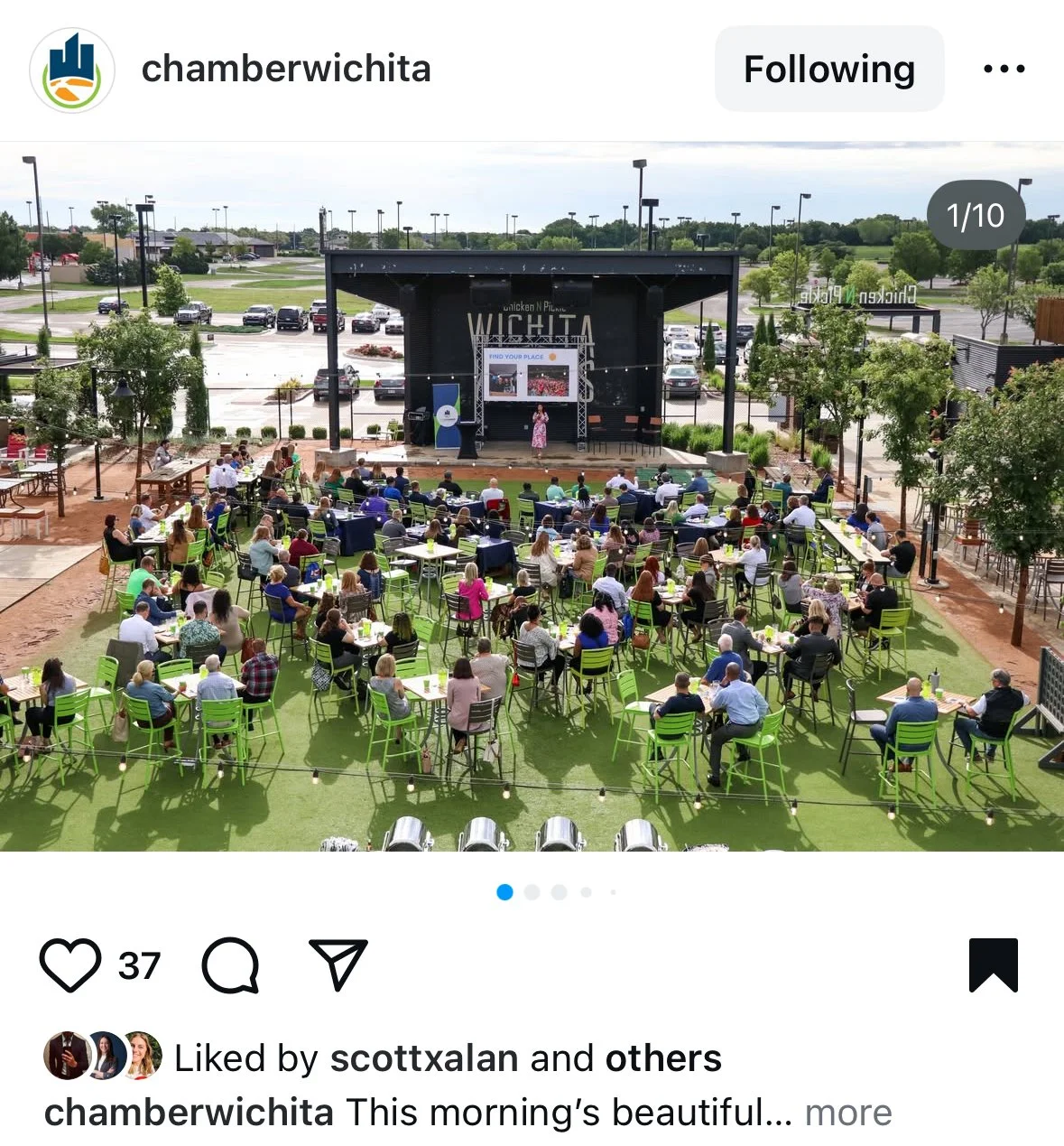 An outdoor event with numerous attendees sitting on green and black chairs at tables, watching a presentation on a stage with large screens, in a park-like setting with trees and a parking lot in the background.