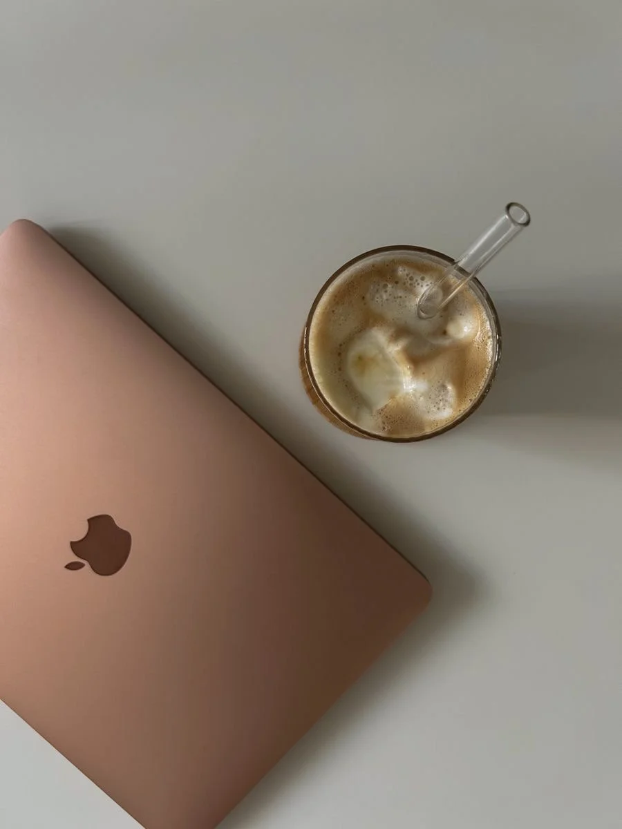 A closed rose gold Apple MacBook computer on a white surface with a glass of iced coffee or frappuccino with foam and a straw beside it.