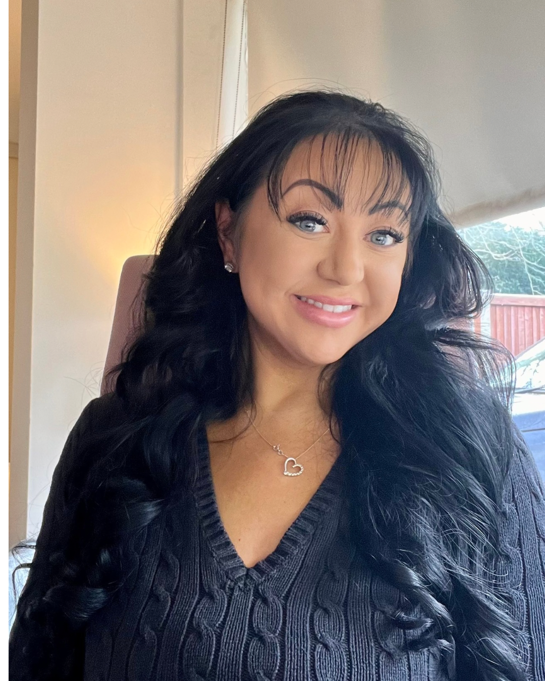 Smiling woman with long black curly hair, wearing a black sweater with a cable knit pattern and a silver heart necklace, sitting indoors near a window.