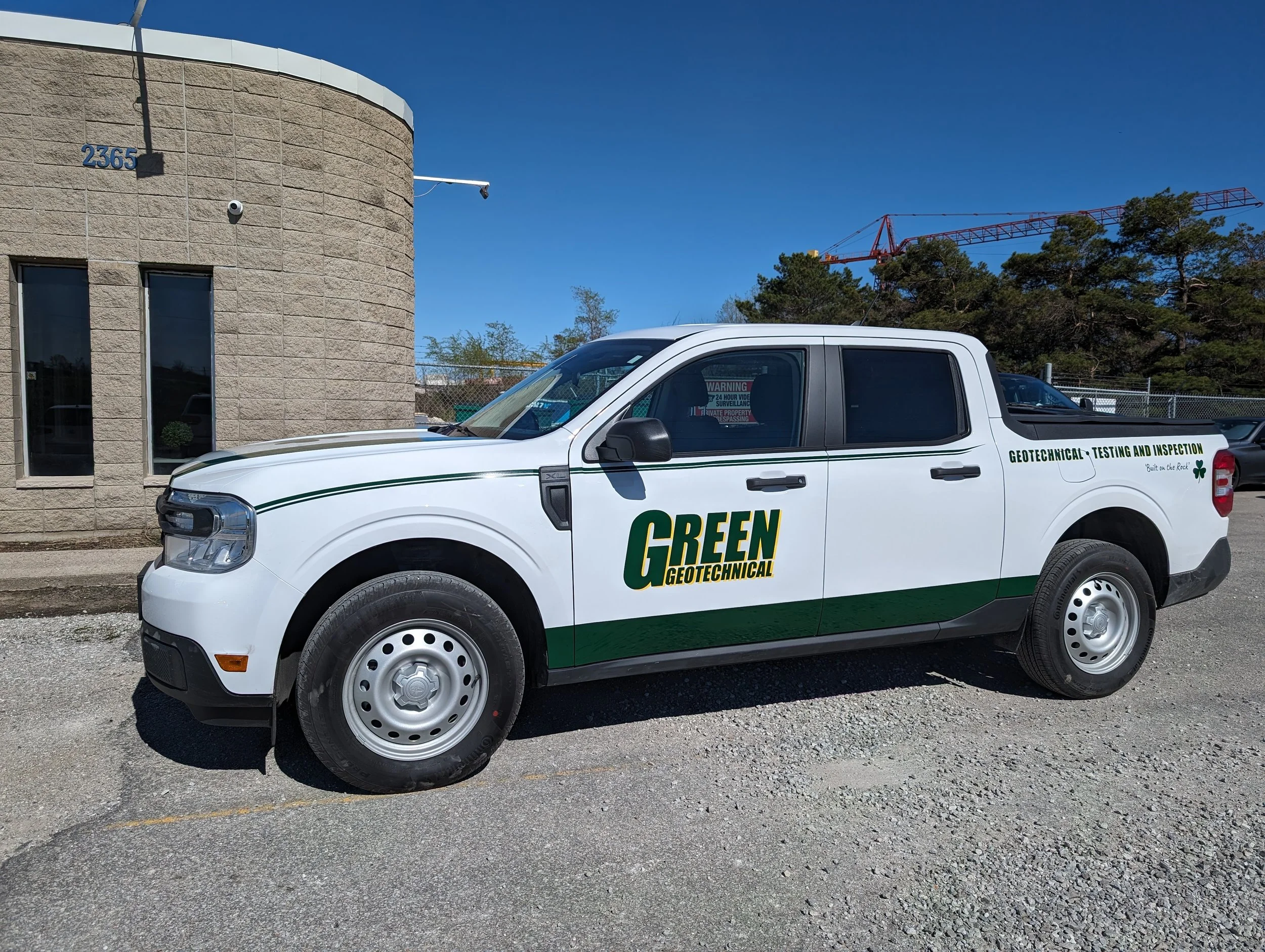 A white pickup truck with green and yellow lettering reads 'Green Geotechnical' and 'Geotechnical Testing and Inspection' parked on a gravel lot. The background includes a beige building, trees, and a construction crane under a clear blue sky.