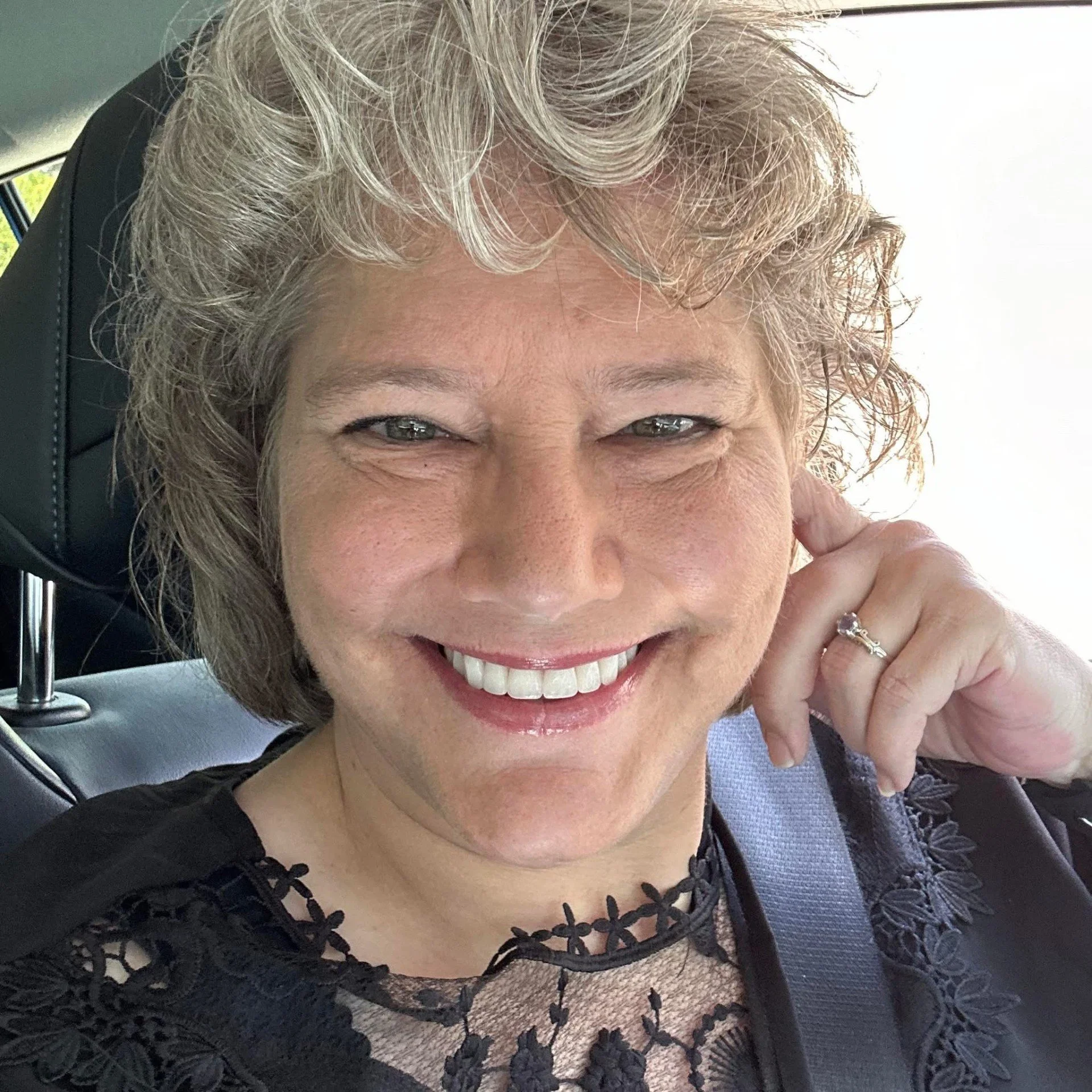Close-up of a smiling woman with short, curly gray hair wearing a black lace top, sitting in a car, showing her ring on her left hand.