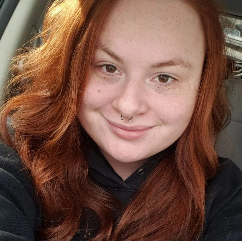 Close-up of a smiling woman with red hair, light skin with freckles, and a septum piercing, wearing a black hoodie, sitting in a car.