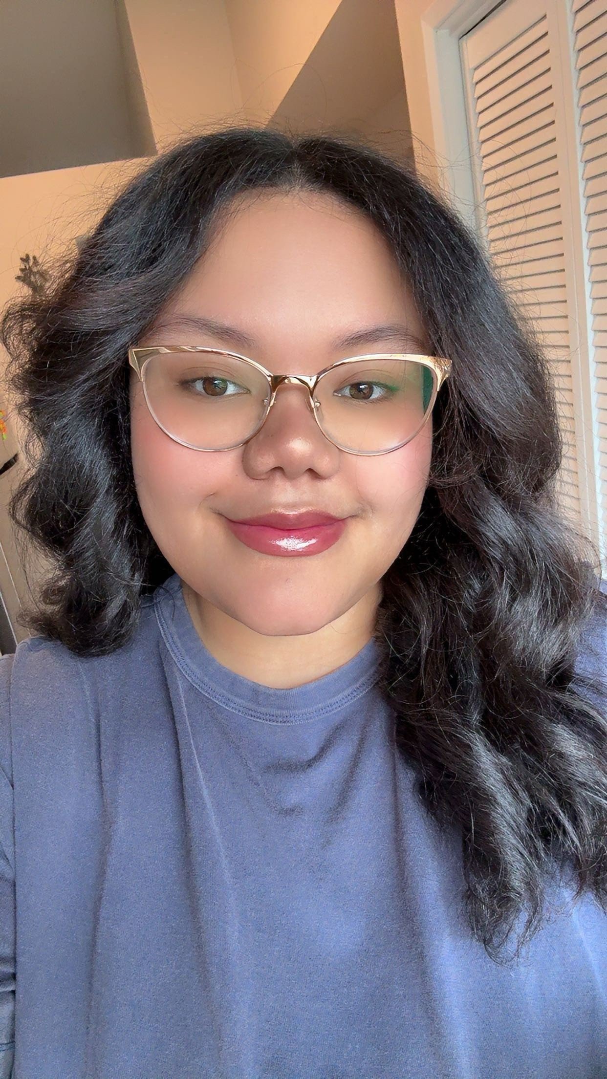 A woman with shoulder-length curly black hair, wearing clear glasses and a blue shirt, smiling in a well-lit room.
