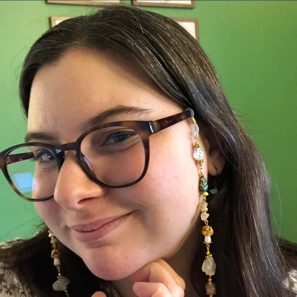 Close-up of a woman with glasses and long dark hair, smiling with her hand under her chin, in front of a green wall with framed pictures.