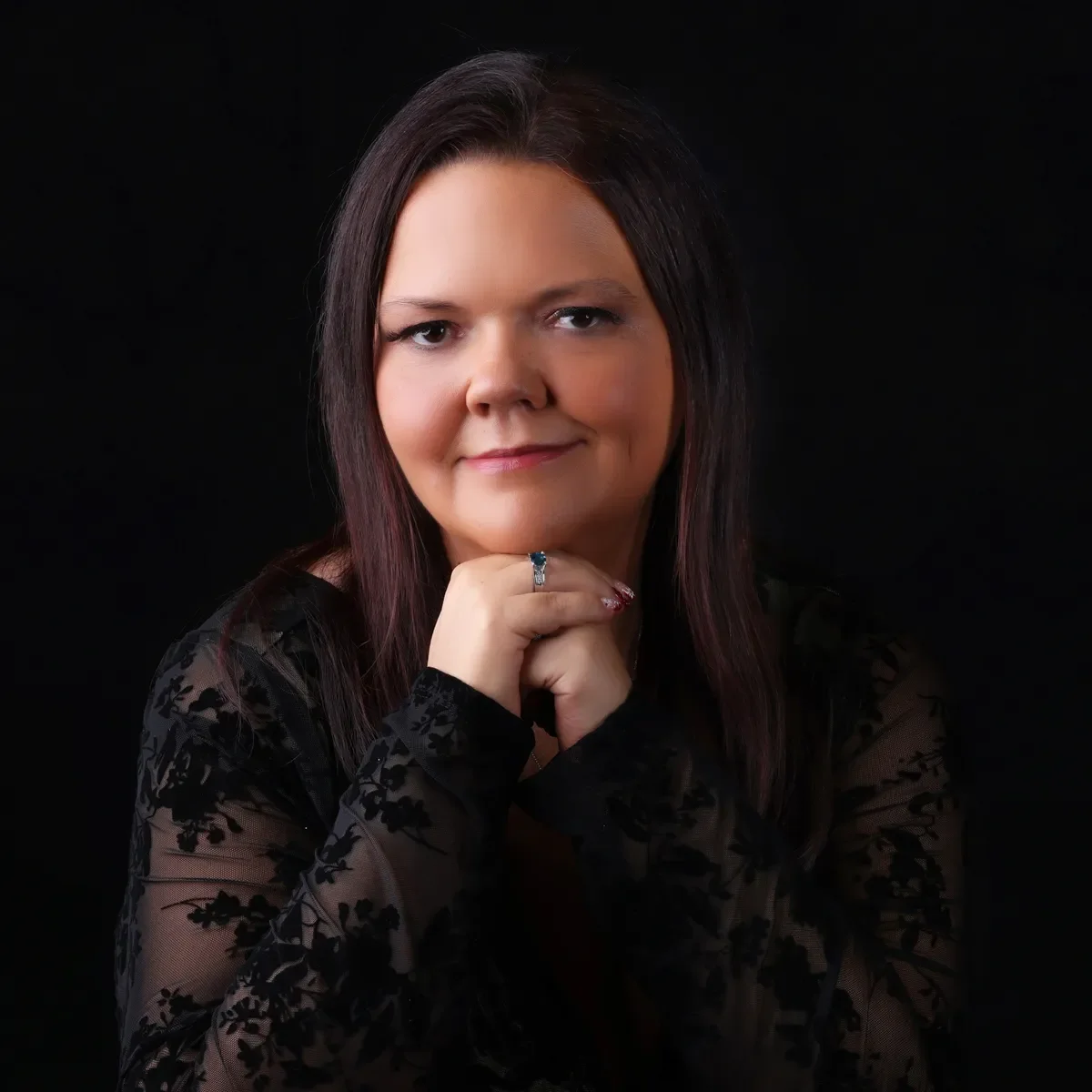 A woman with shoulder-length dark hair, wearing a black lace top, resting her chin on her hand with a confident smile, against a solid black background.