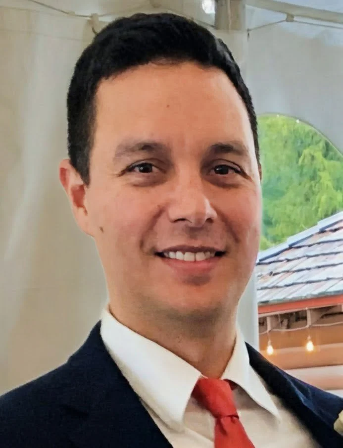 A man with short black hair, wearing a dark suit, white shirt, and red tie, smiling at the camera with a blurred outdoor background.