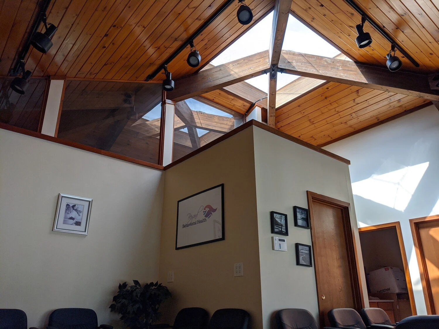 Interior of a room with a high wooden ceiling, skylights, track lighting, and a small seating area with chairs. There are framed pictures on the wall, a potted plant, and a doorway leading to another room.
