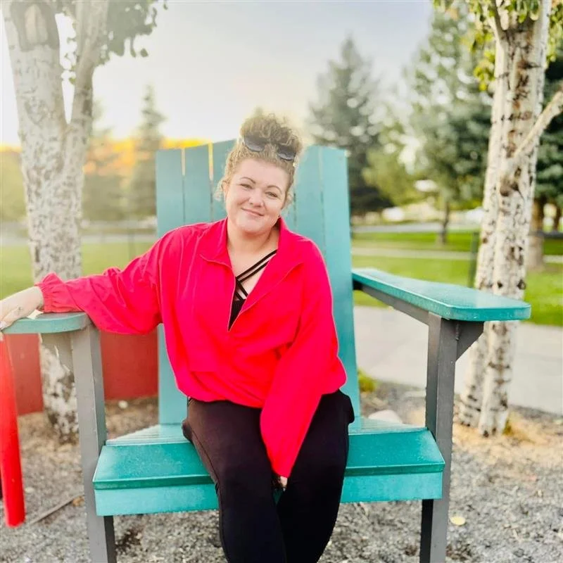 A woman sitting on a large turquoise wooden chair outdoors in a park, with trees and a grassy area in the background, during what appears to be late afternoon or early evening.