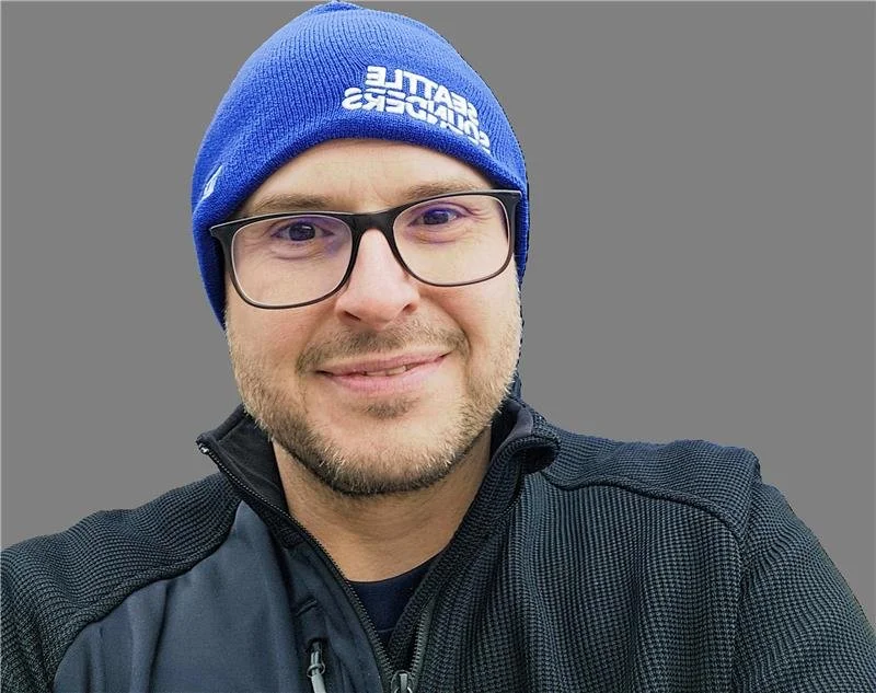 A man wearing glasses, a blue knit hat with white text, and a black zip-up jacket, smiling in front of a plain gray background.