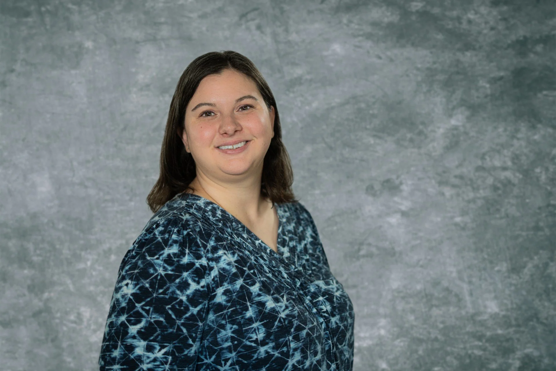 Young woman with dark hair standing infront of a marble gray background with a smile.
