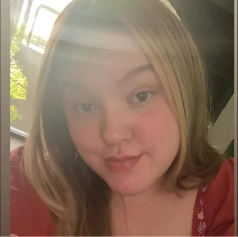 A young woman with blonde hair taking a selfie indoors with sunlight coming through a window behind her.