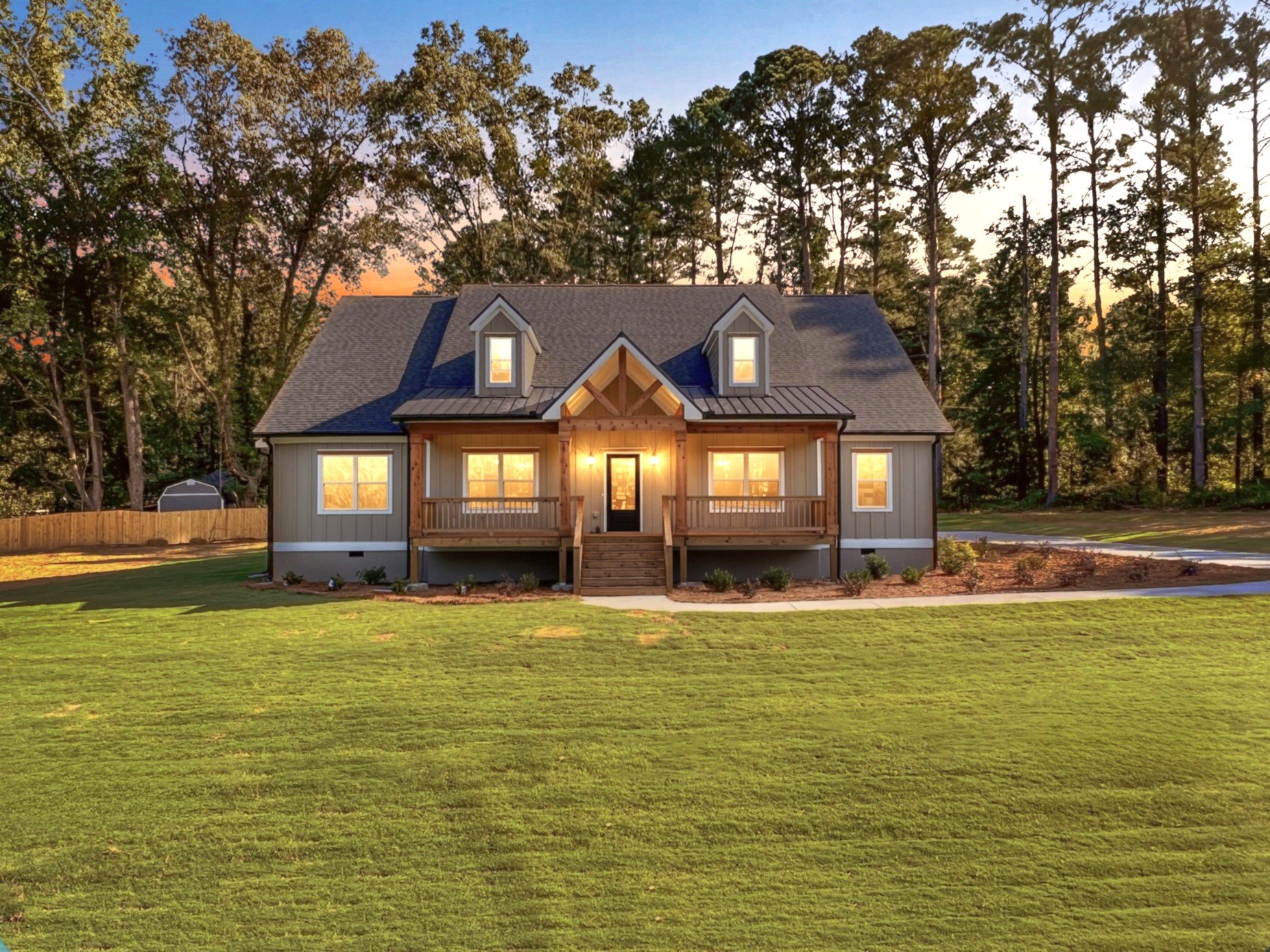 A newly built house with a front porch, illuminated windows, surrounded by a well-maintained lawn and tall trees at dusk.