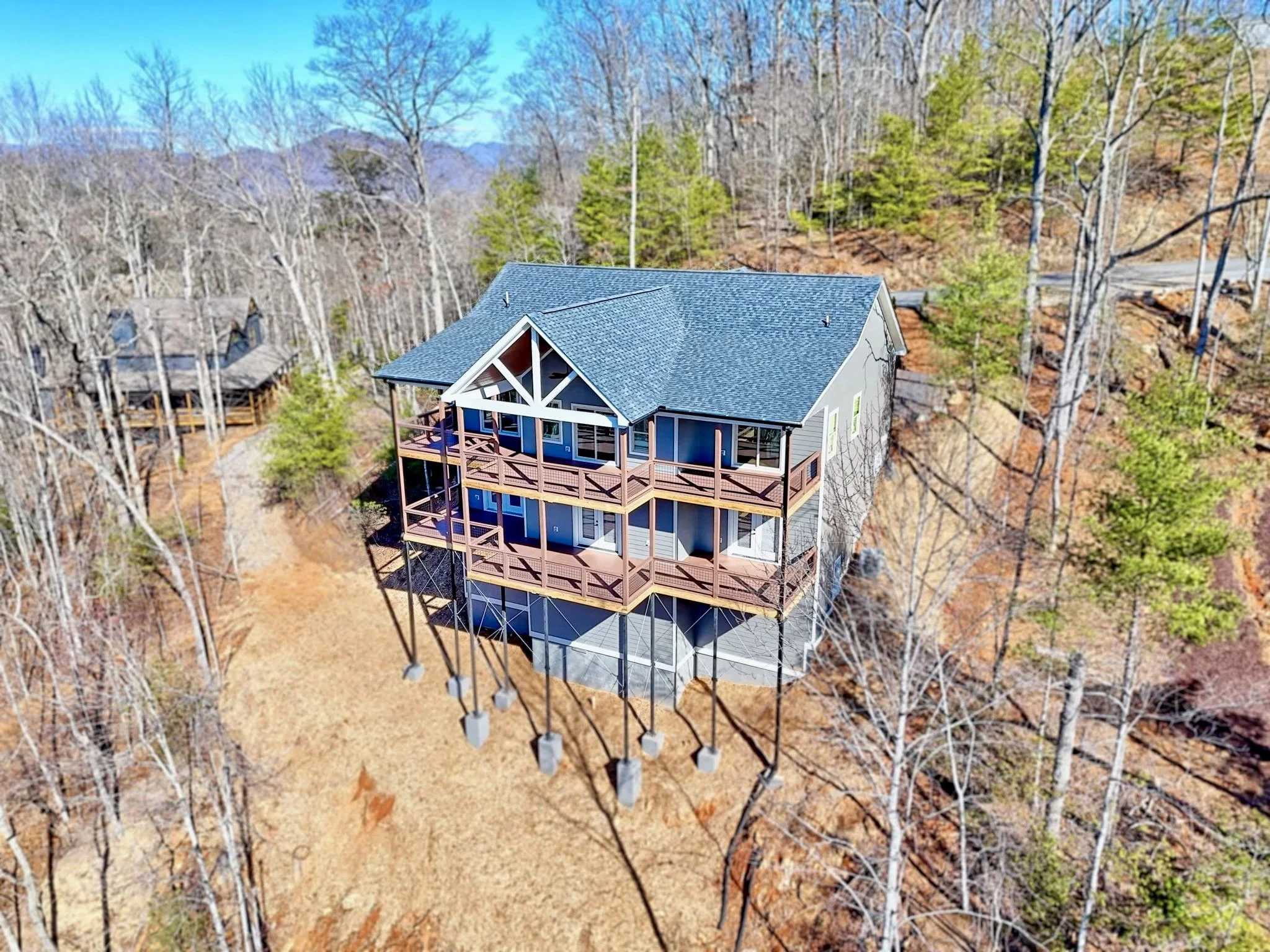 A multi-story house with a porch, located in a wooded area with tall, leafless trees, and a partly visible road in the background.