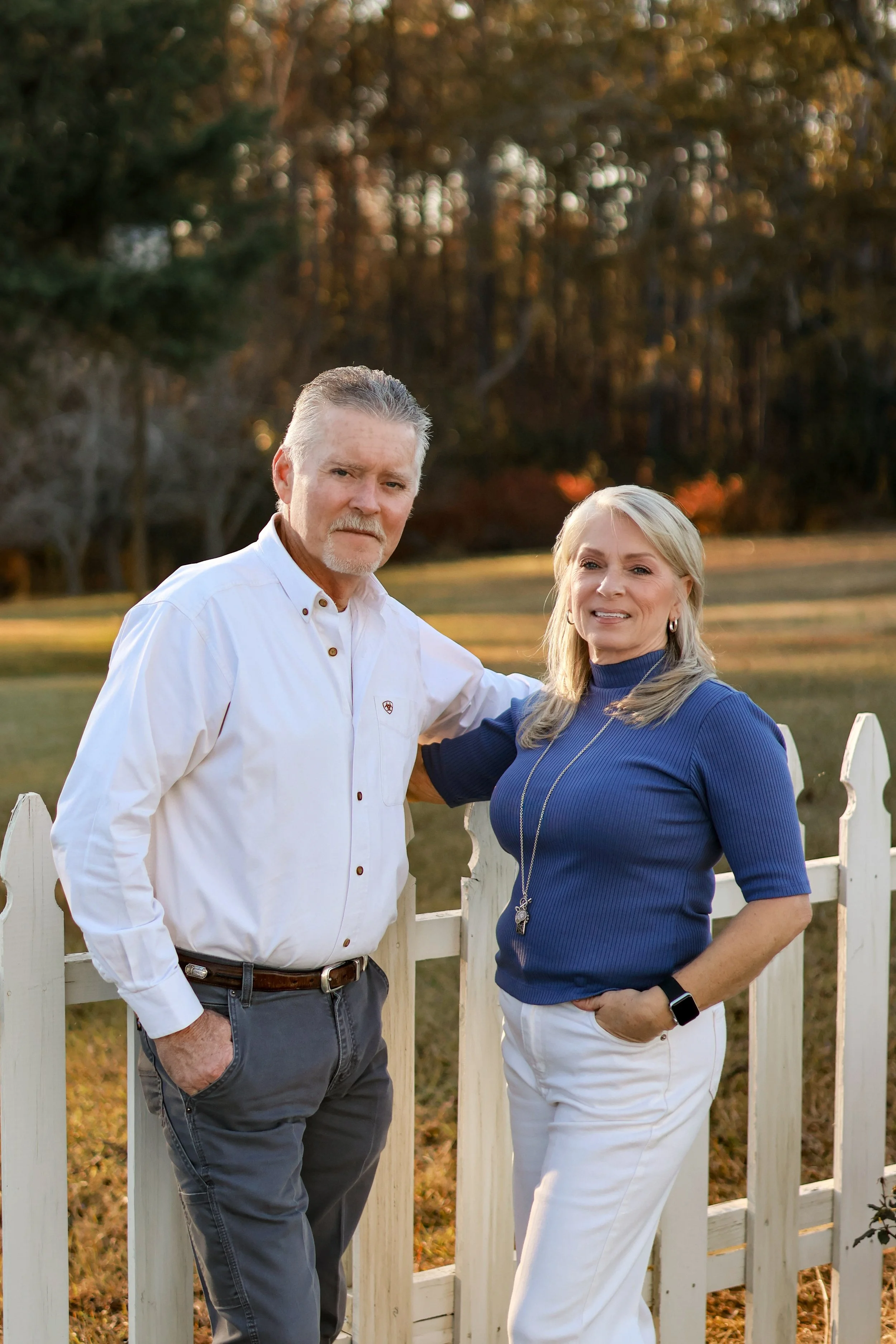 A middle-aged man and woman standing outdoors near a white picket fence during sunset, with trees in the background. The man is wearing a white button-up shirt and gray pants, and the woman is wearing a blue top and white pants. She has her hand in her pocket and is wearing a necklace and smartwatch.
