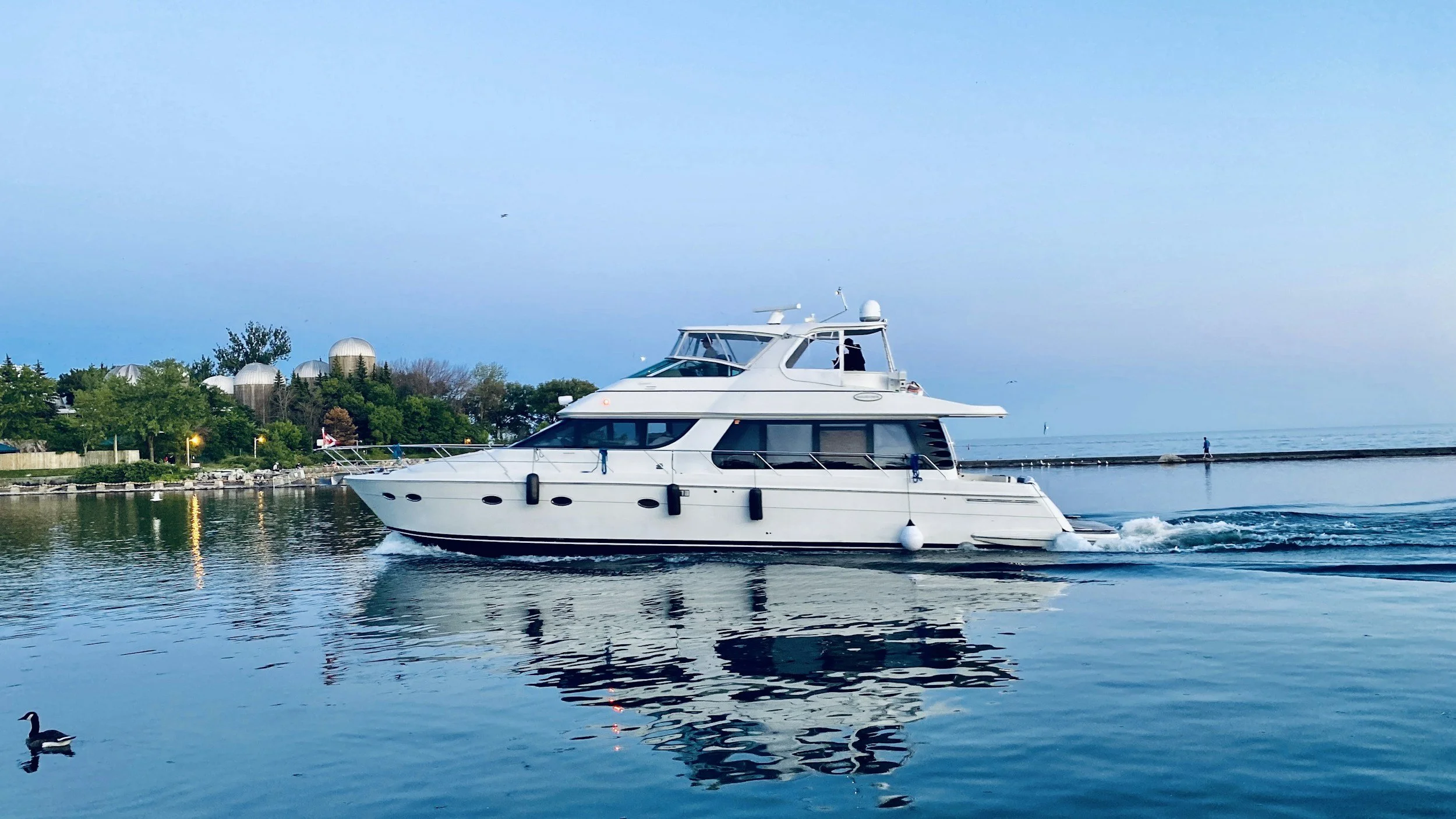 A white yacht cruising on a calm body of water near a shoreline with trees and dome structures, under a clear blue sky, with a person walking on the pier in the distance. This could be owned by Jeff Bezos soon.