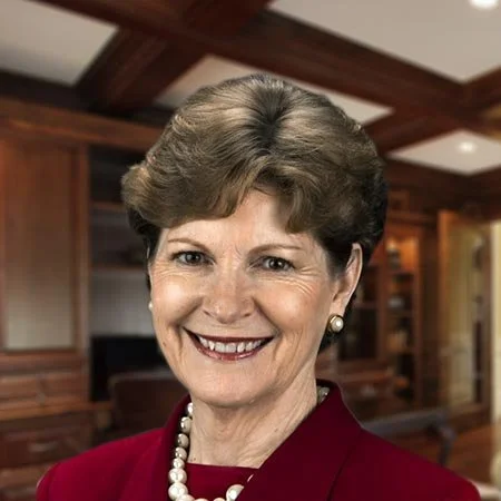 A smiling elderly woman with short brown hair, wearing earrings, a pearl necklace, and a red blazer, indoors with wooden paneling and furniture in the background.