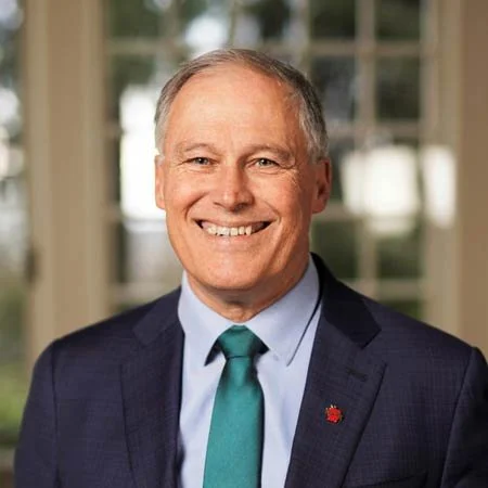 A smiling middle-aged man wearing a dark suit, light blue shirt, and teal tie, standing indoors in front of windows.