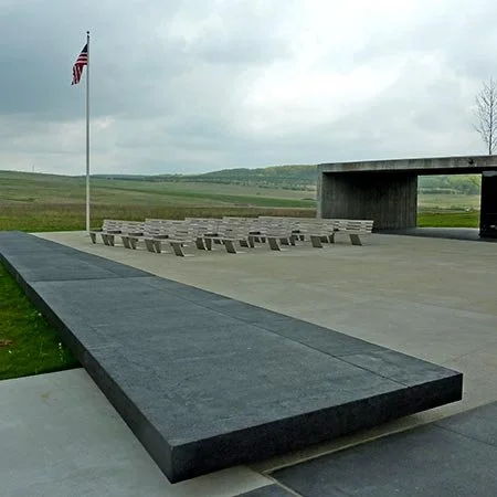 A memorial site with a flagpole flying the American flag, benches, and a concrete structure, set in a grassy open field under cloudy skies.