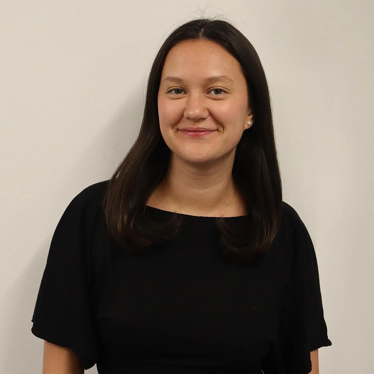 A young woman with dark brown hair, wearing a black top, smiling, standing against a plain white wall.