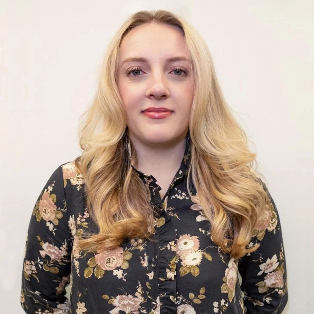 A woman with long, wavy blonde hair and a floral black blouse standing against a plain white wall.
