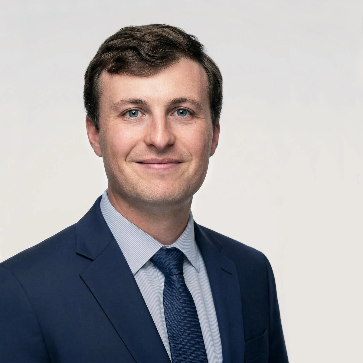Headshot of a man with brown hair and blue eyes, wearing a dark suit, light blue shirt, and matching tie, against a plain light background.