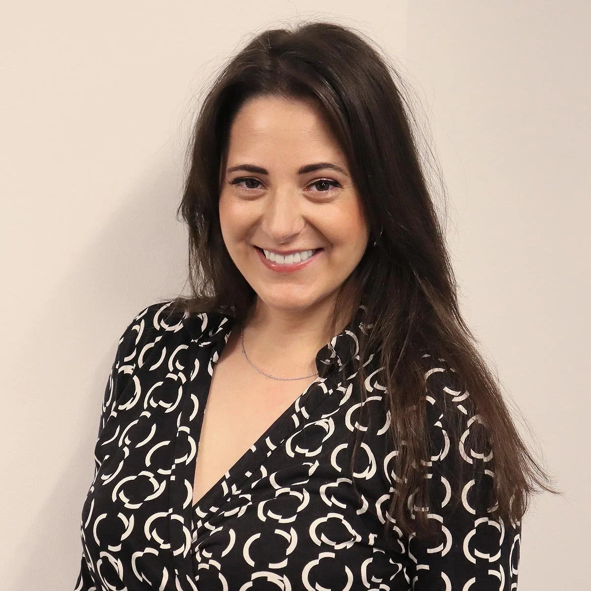 A woman with long dark hair, smiling, wearing a black and white patterned blouse and a necklace, standing against a plain wall.