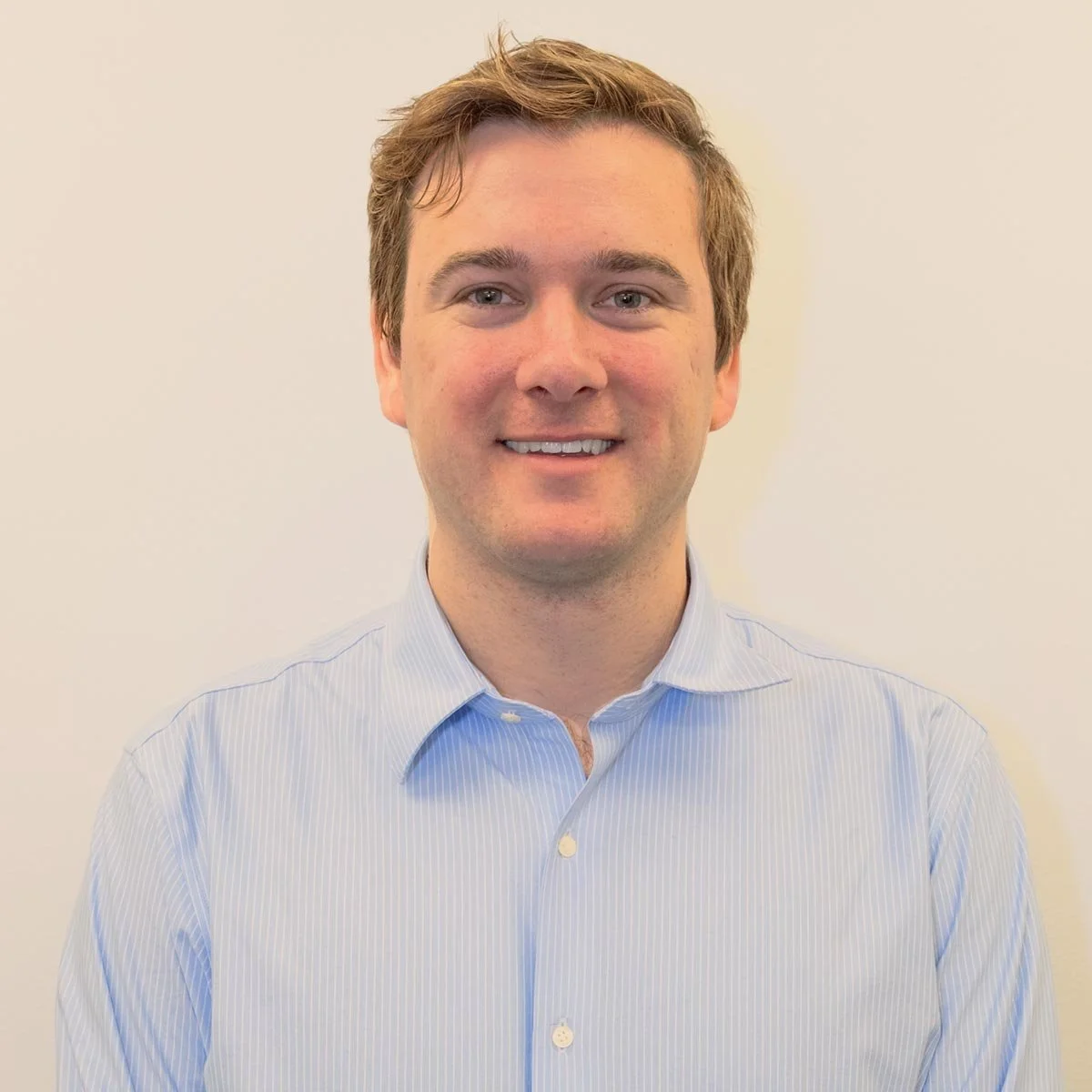 Headshot of a young man with light brown hair smiling, wearing a light blue striped dress shirt, against a plain off-white background.