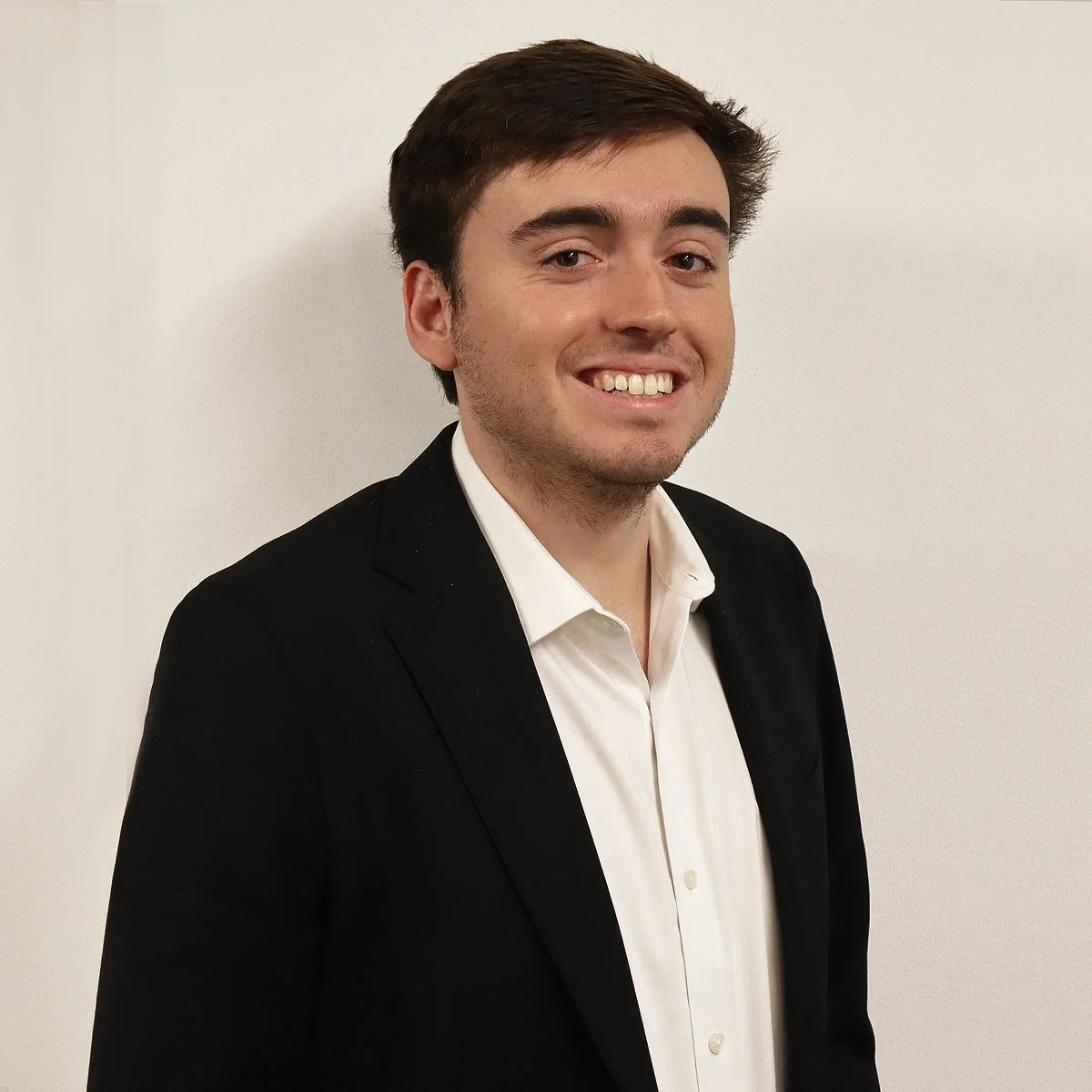A young man with brown hair, wearing a white shirt and a black blazer, smiling and standing against a plain white wall.
